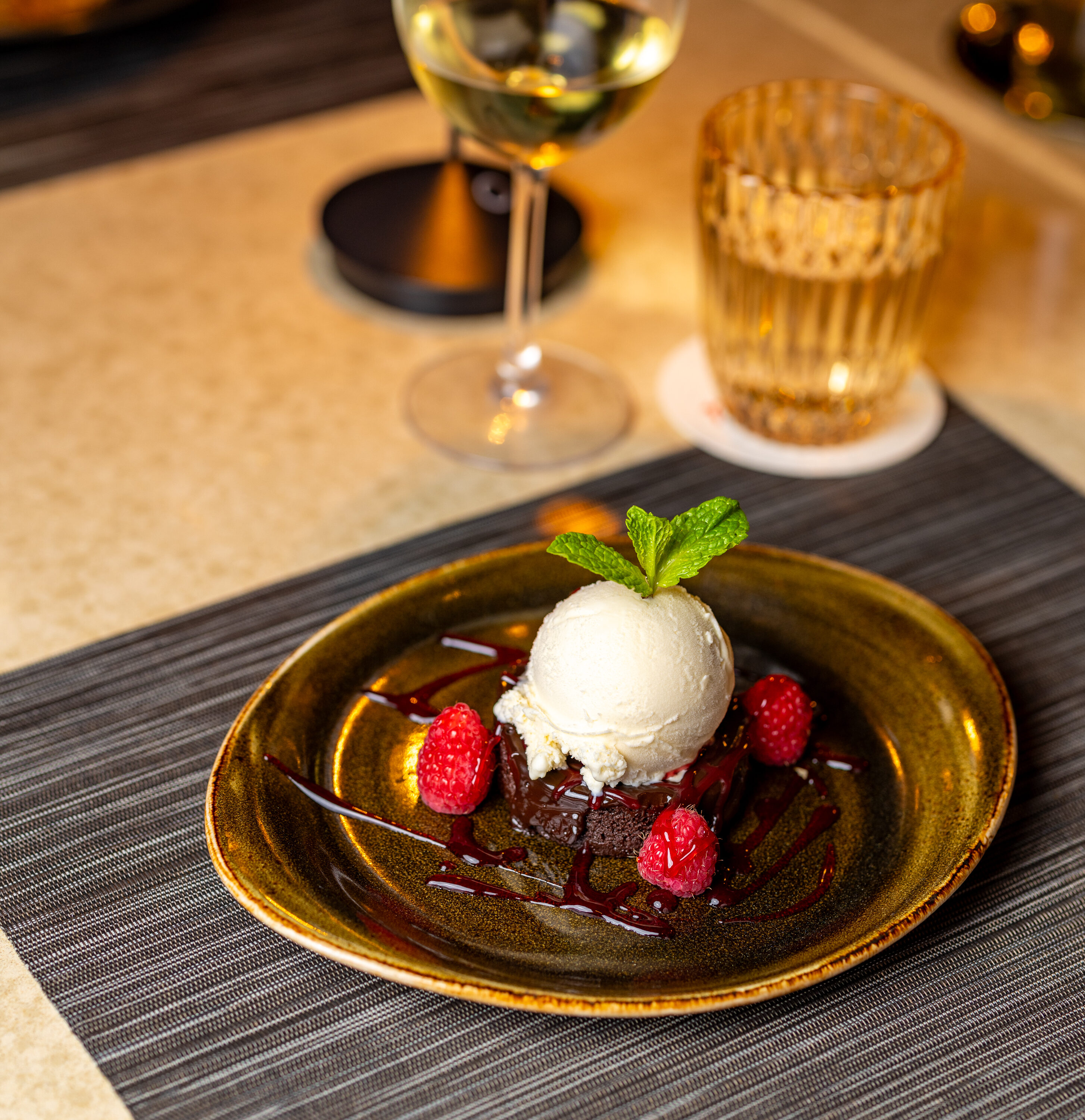 A dessert plate with a brownie topped with a scoop of vanilla ice cream, raspberries, mint, and sauce, set next to a glass of white wine and a glass of water on a table.