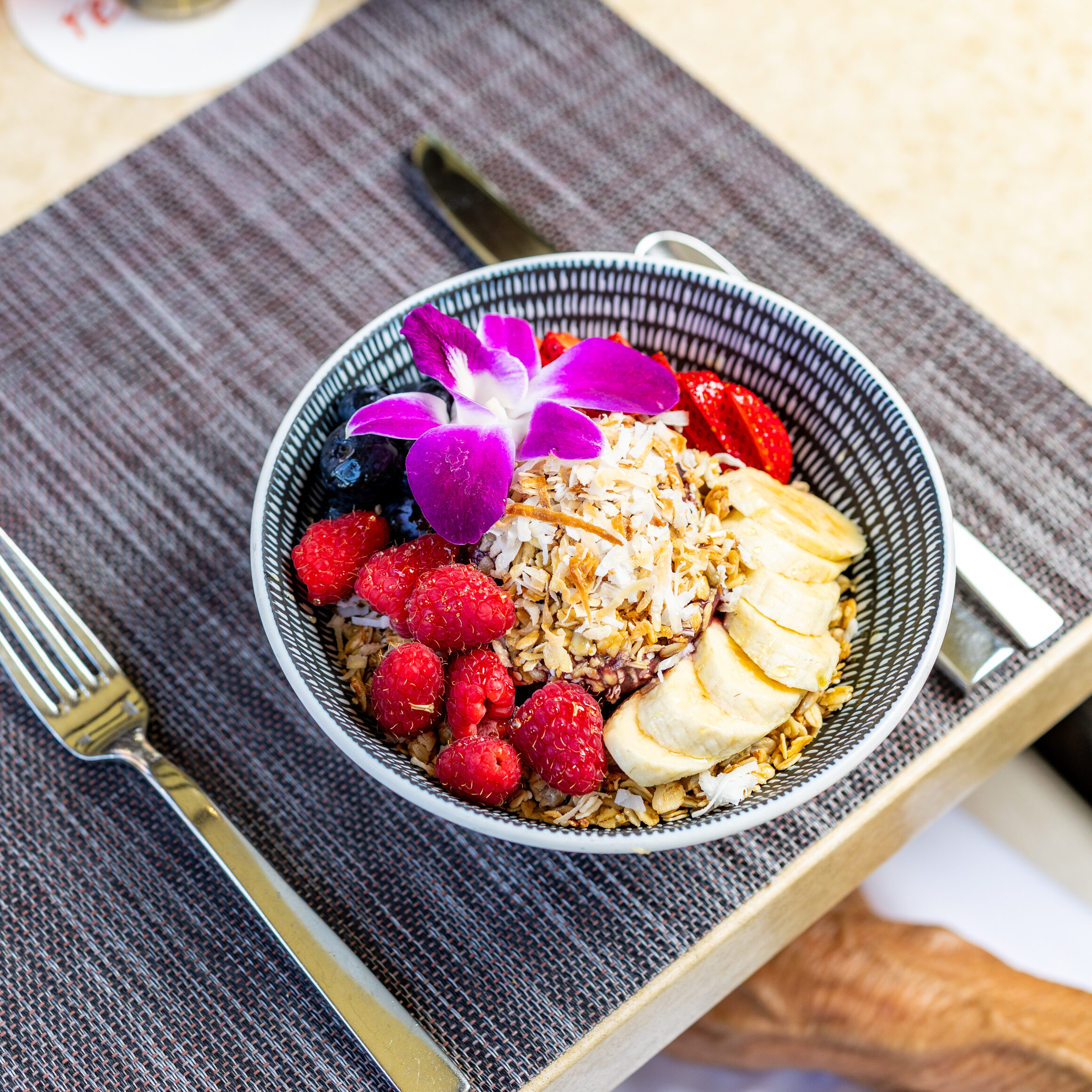 A bowl of granola topped with raspberries, banana slices, strawberries, coconut, and a purple orchid sits on a placemat next to a fork, knife, and two drinks—perfect for special celebrations.