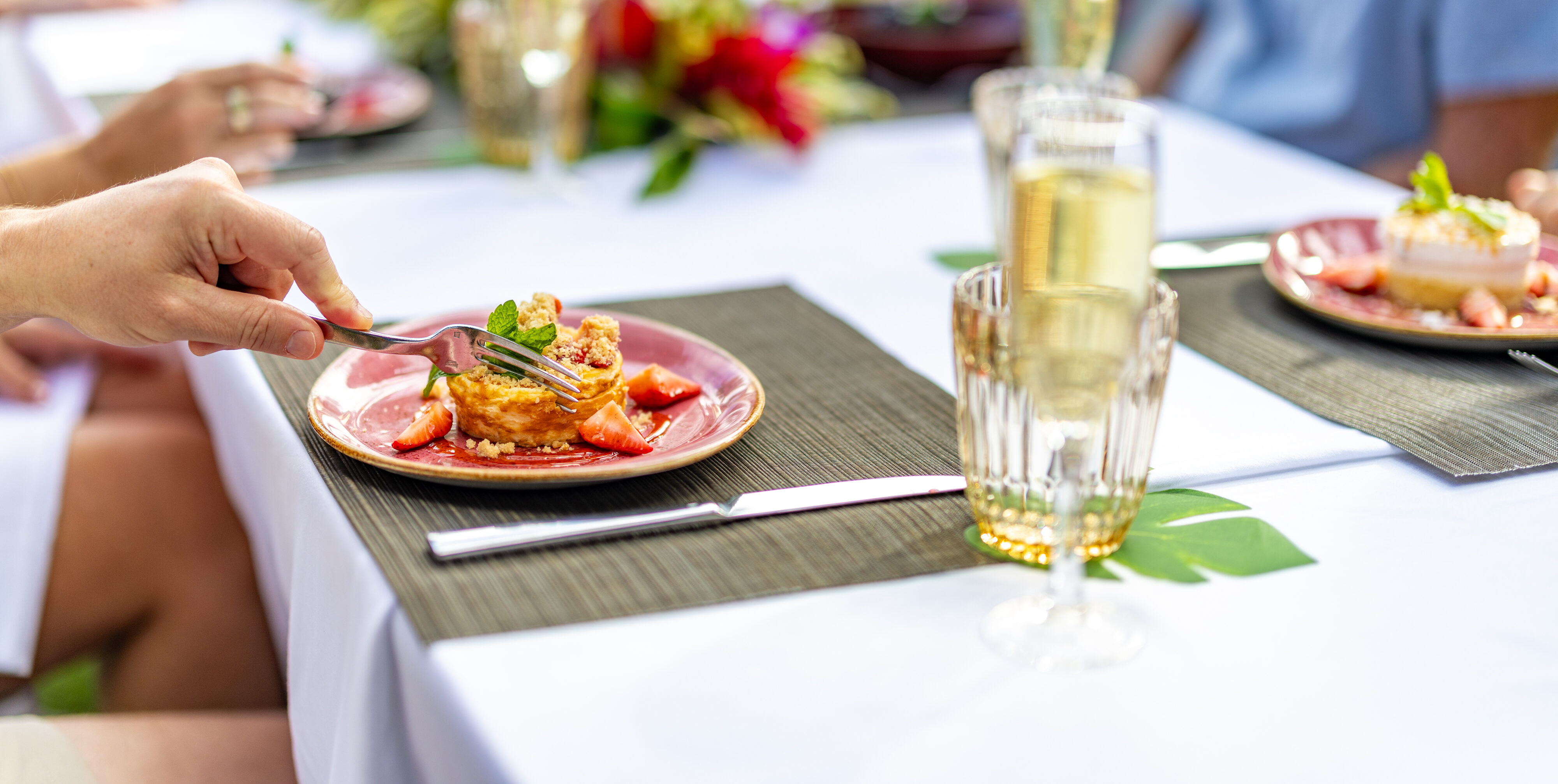 A person uses a fork to eat a plated dessert with strawberries at an outdoor table set with drinks and flowers.