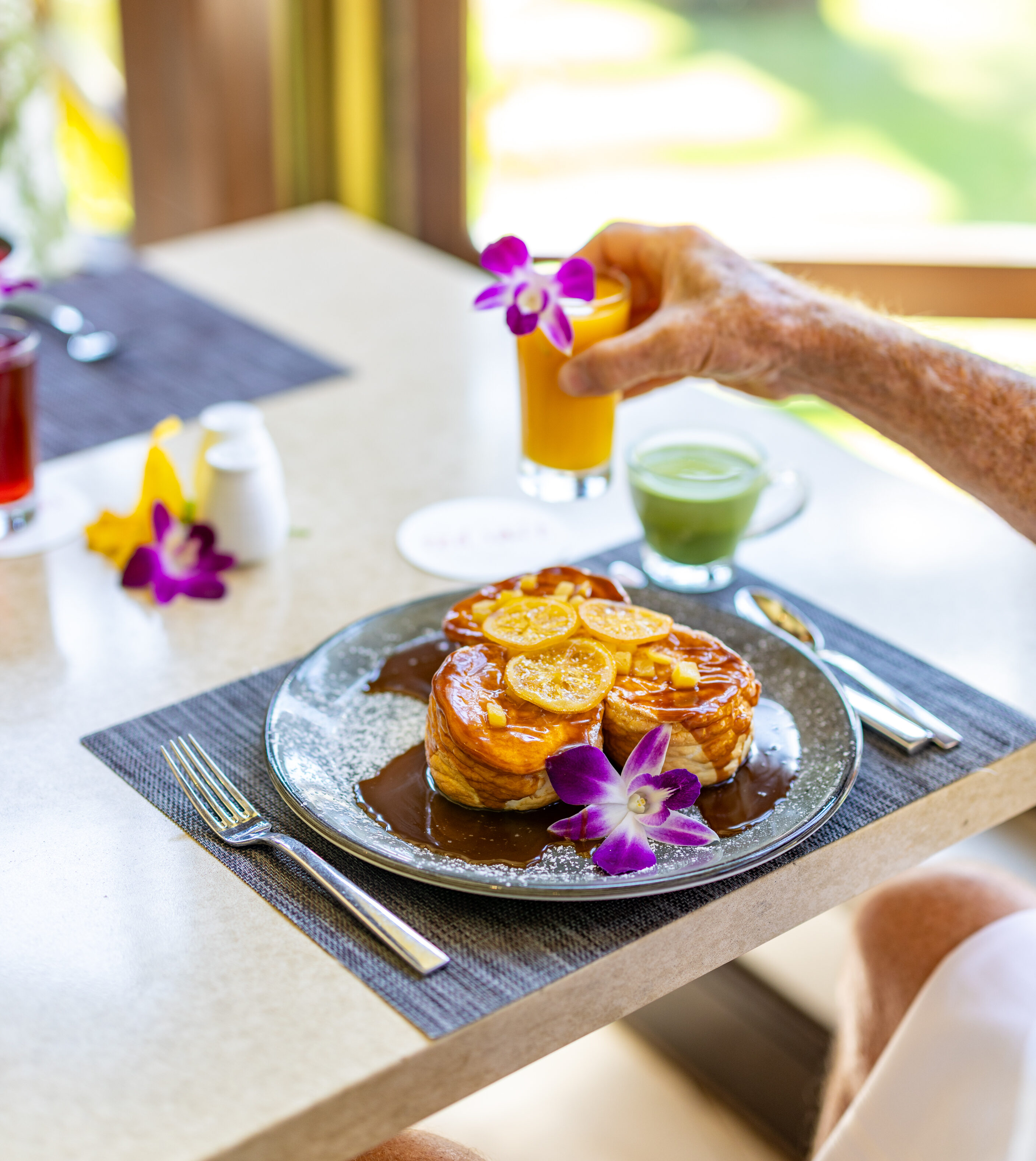 A plate of pancakes with orange slices and purple orchids, next to a green drink; a person reaches for an orange beverage on a sunlit table.