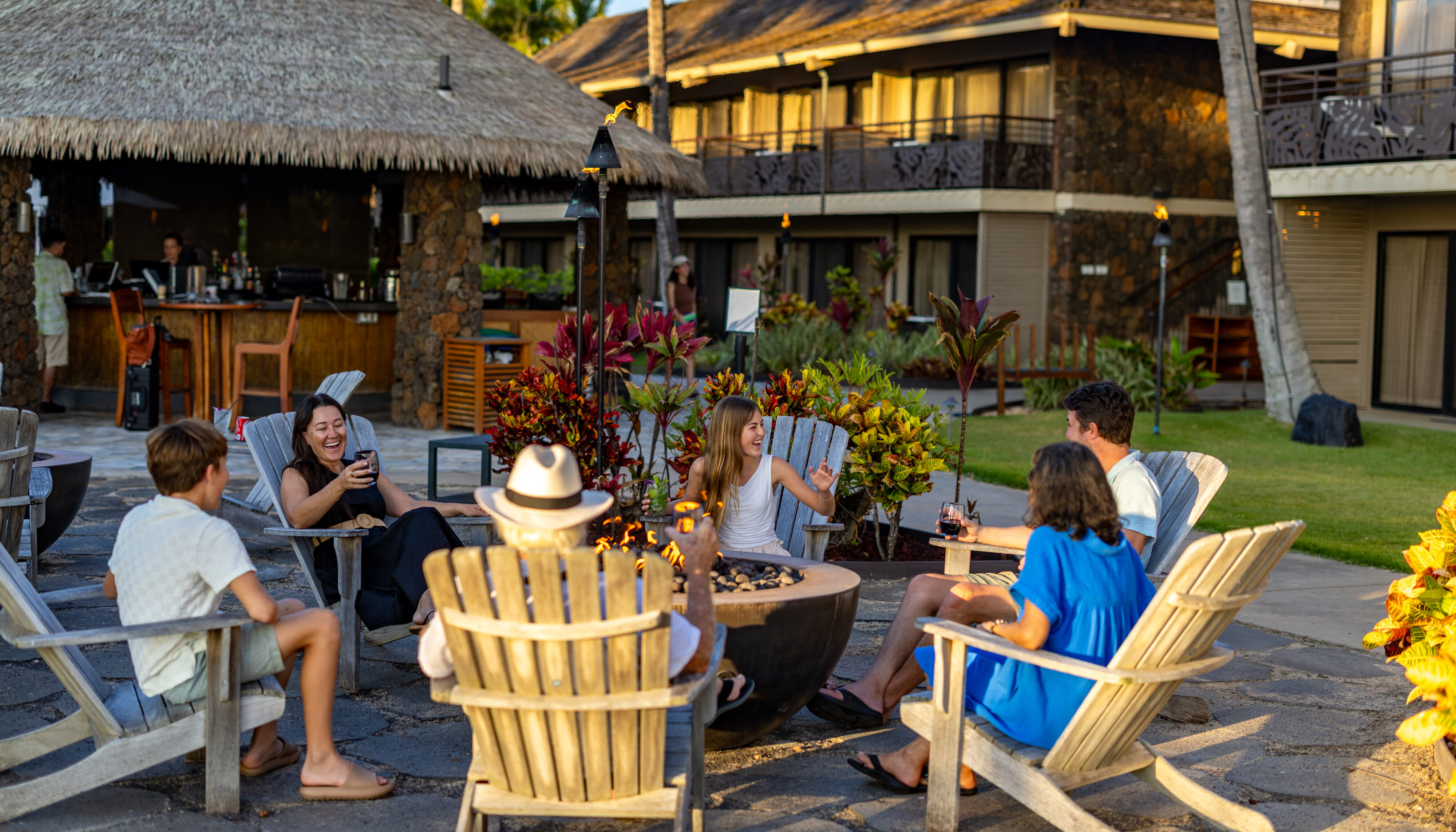 A group of people sit around a fire pit in wooden chairs, talking and enjoying drinks at an outdoor resort area with tropical plants and buildings in the background.