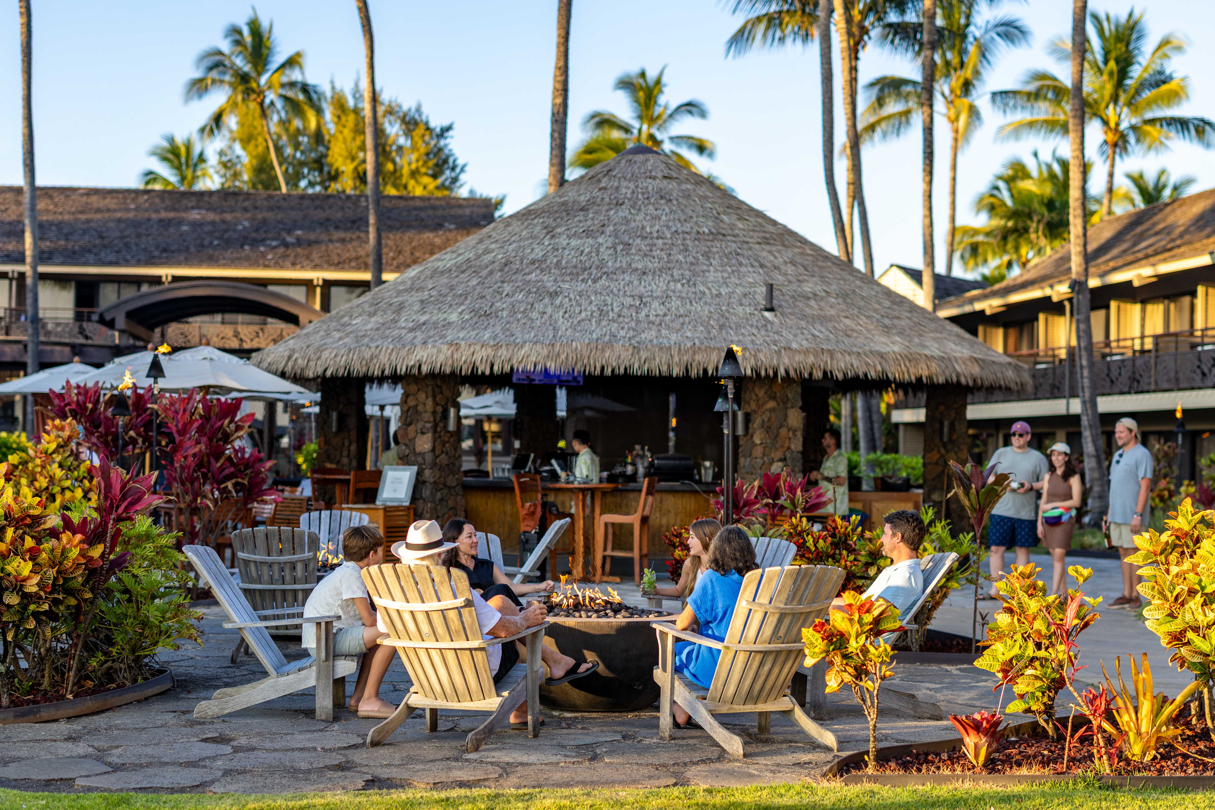 People sit around a fire pit on wooden chairs at an outdoor resort area with tropical plants, a thatched-roof hut, and palm trees in the background.