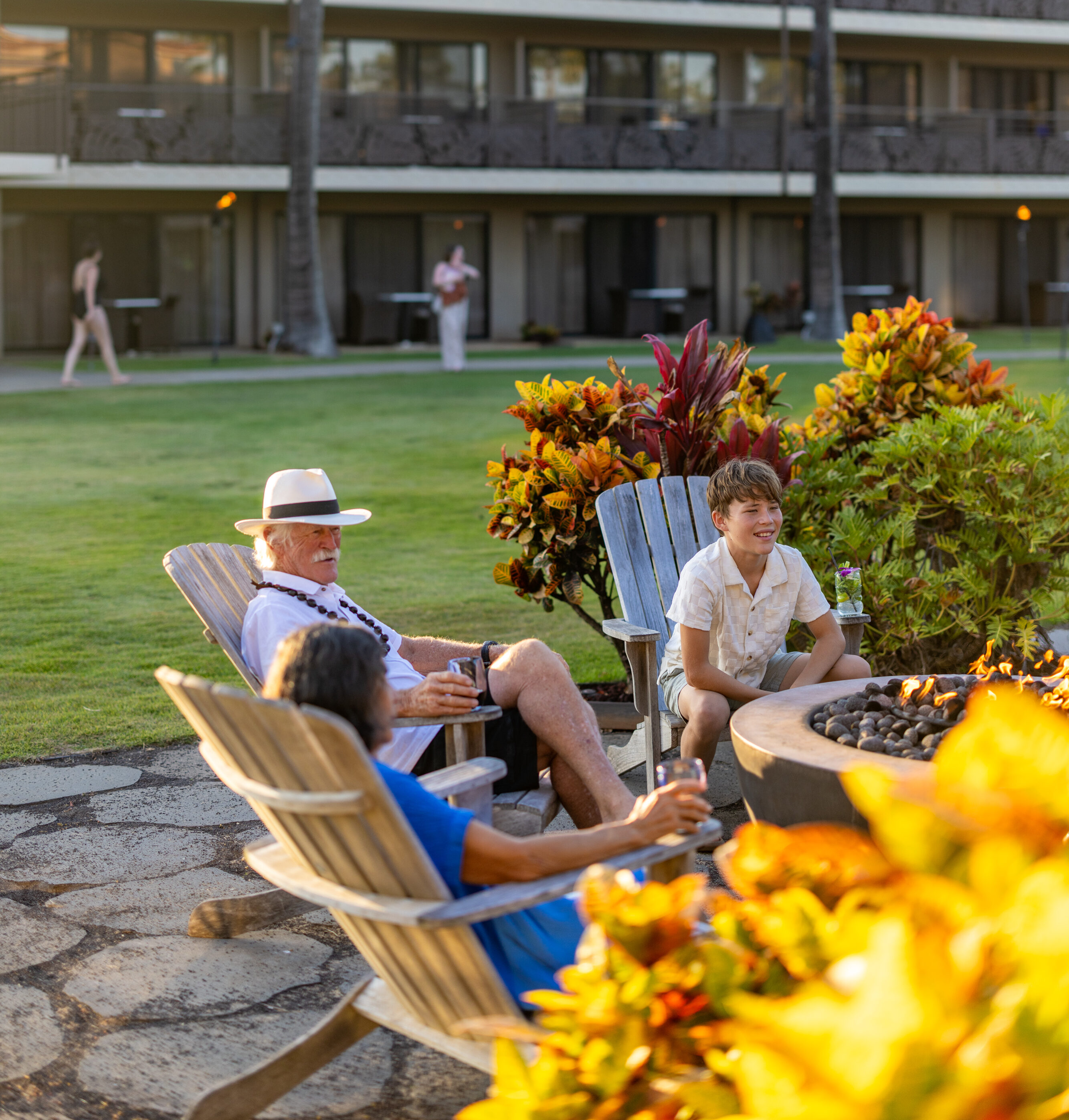 Three people sit in Adirondack chairs around a fire pit on a lawn with a resort building and palm trees in the background.