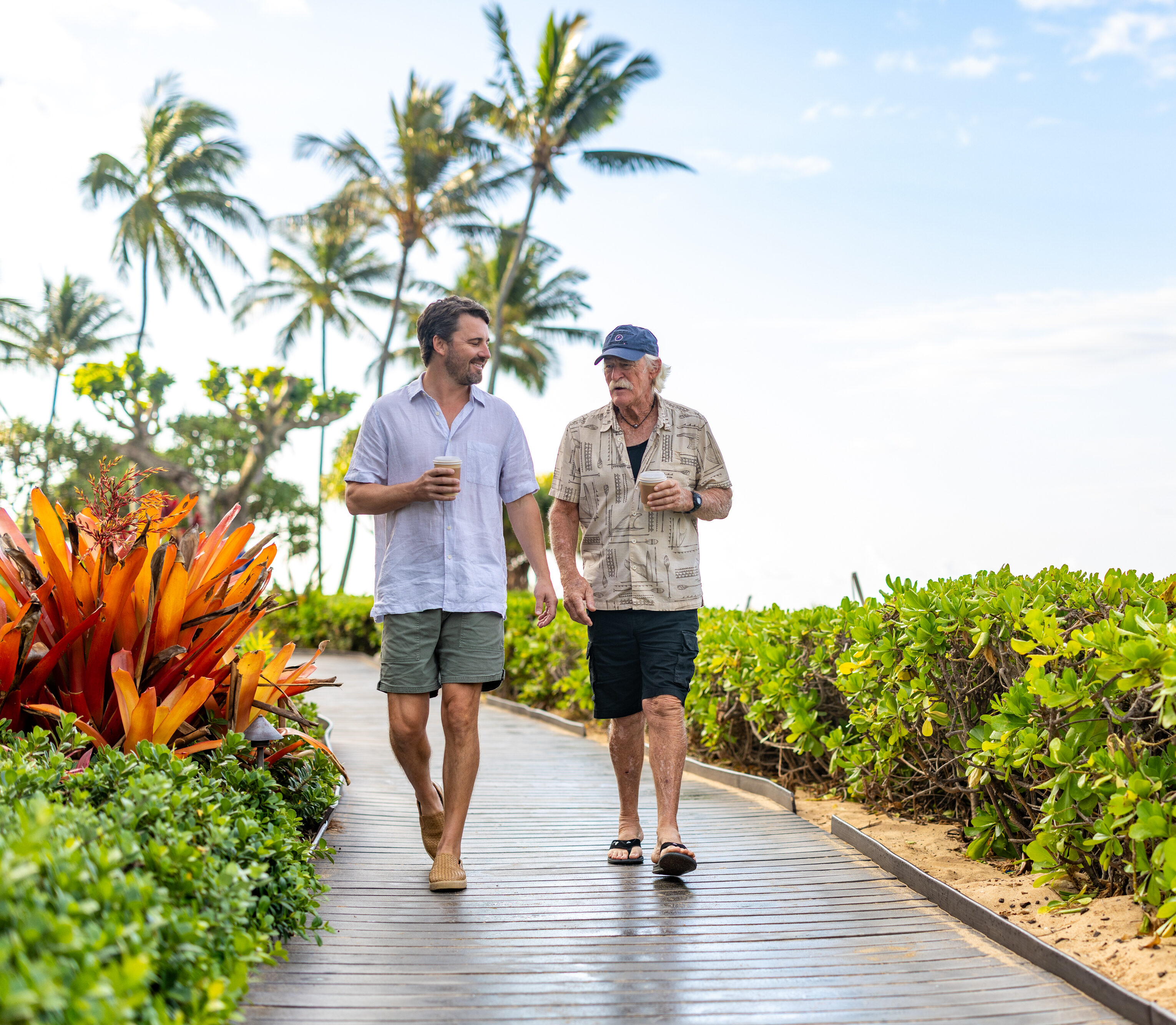 Two men walking on a boardwalk by the beach, holding drinks and talking, with tropical plants and palm trees in the background.