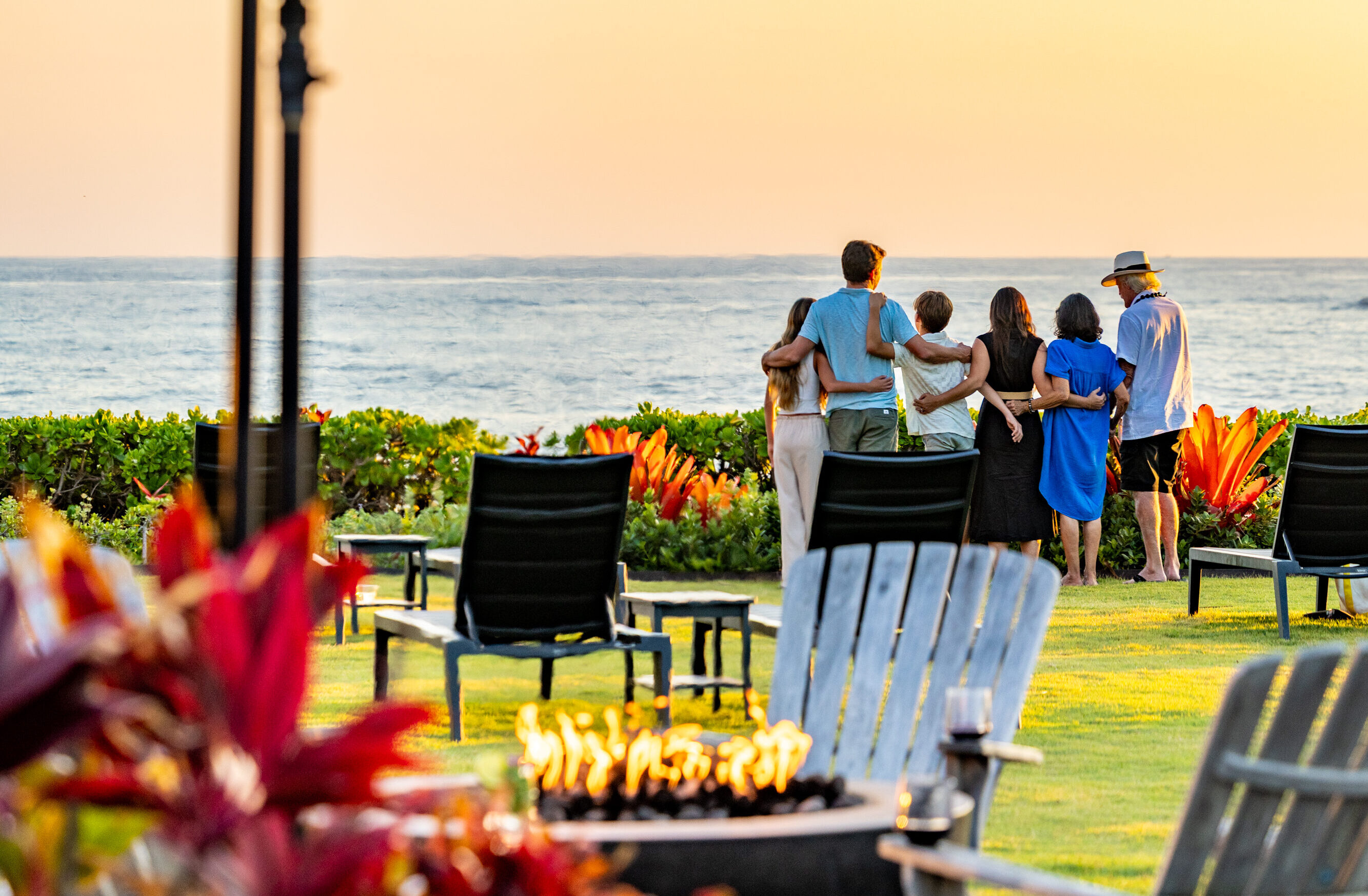Five people stand together facing the ocean at sunset, surrounded by lawn chairs, plants, a lit fire pit, and a tiki torch in the foreground—capturing the warmth of gatherings and celebrations.