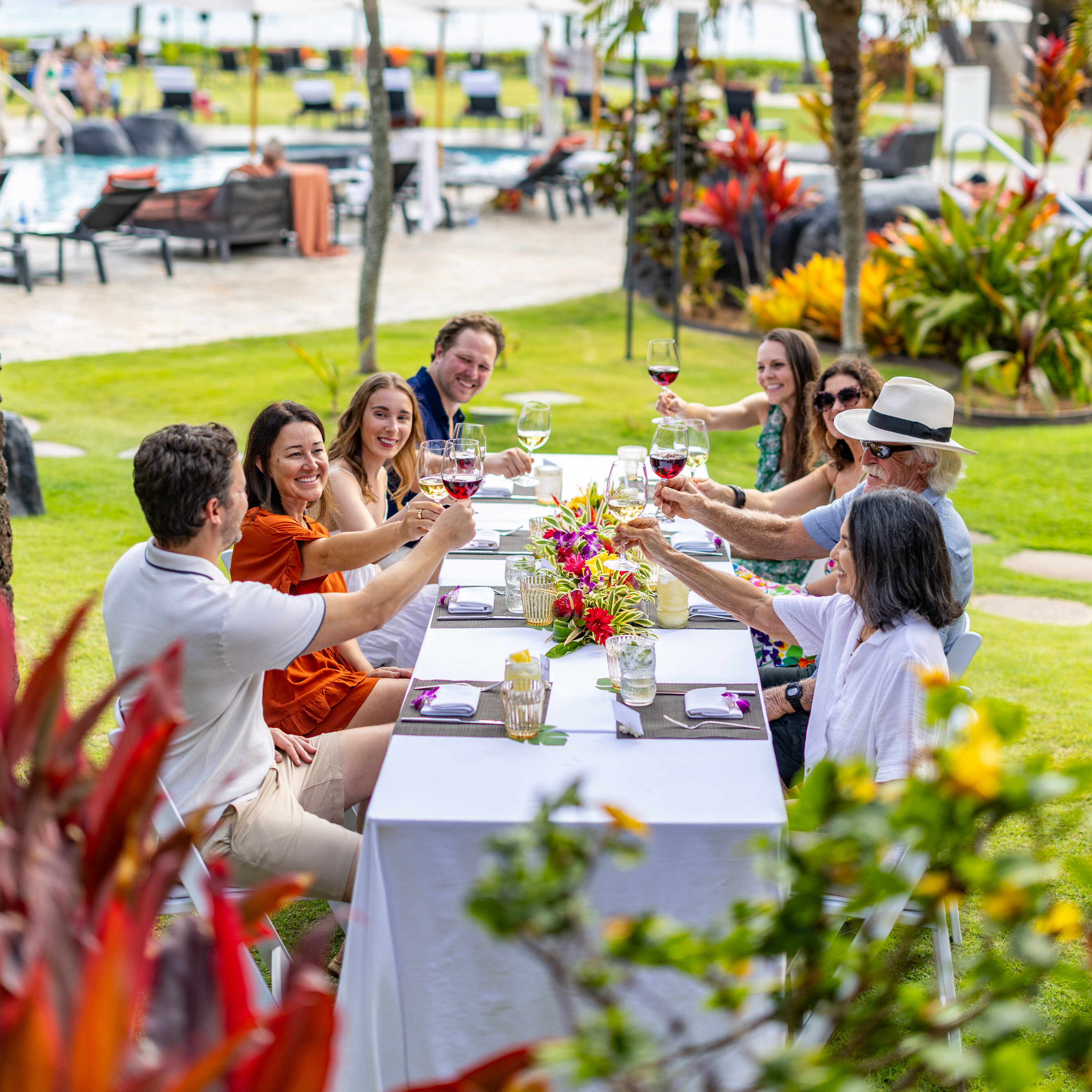 A group of people sit at a long outdoor table, raising glasses in a toast, surrounded by tropical plants and palm trees near a pool.