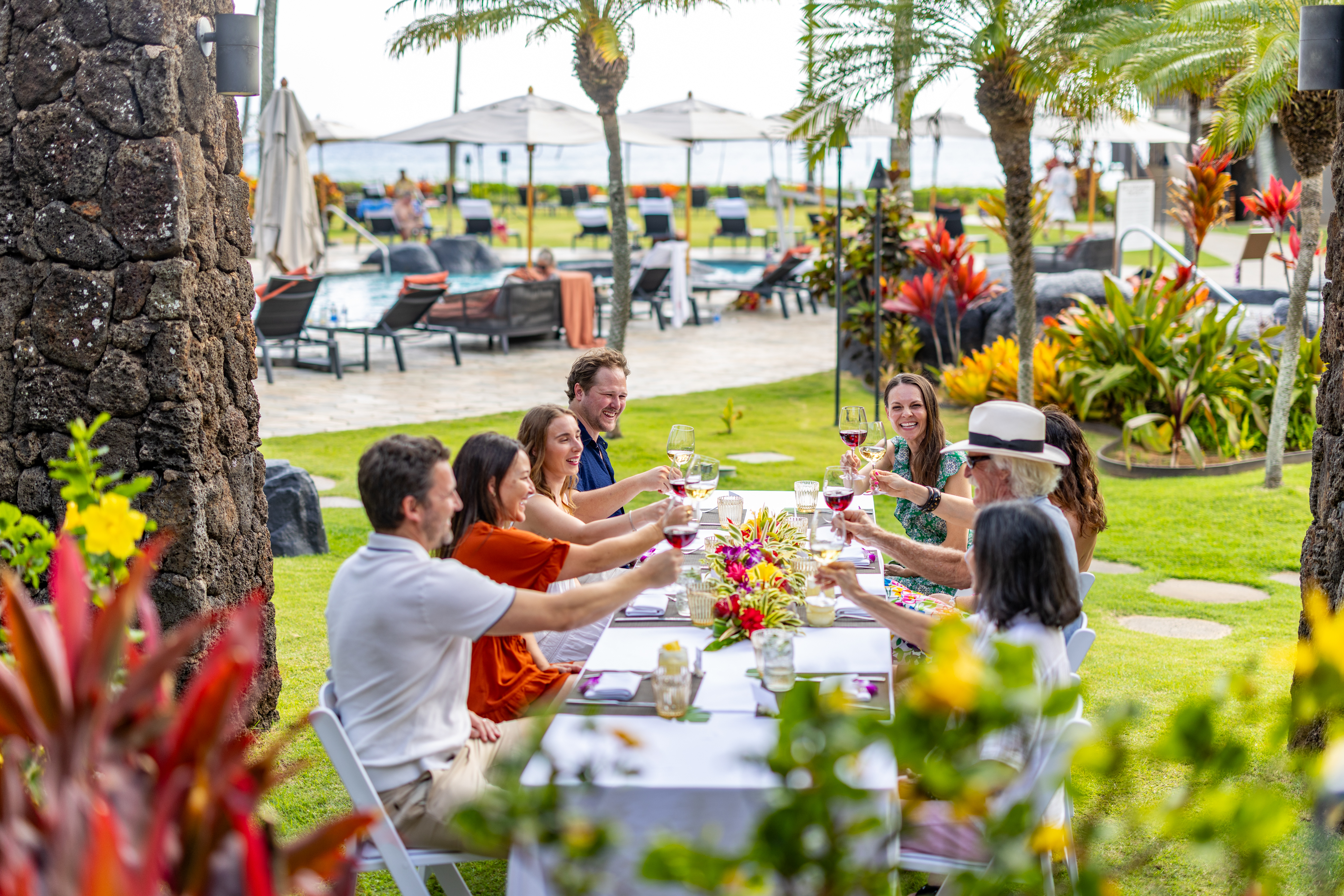 A group of people sit at an outdoor table, raising glasses in a toast during a lively Paina Dinners gathering, surrounded by tropical plants and poolside lounge chairs.