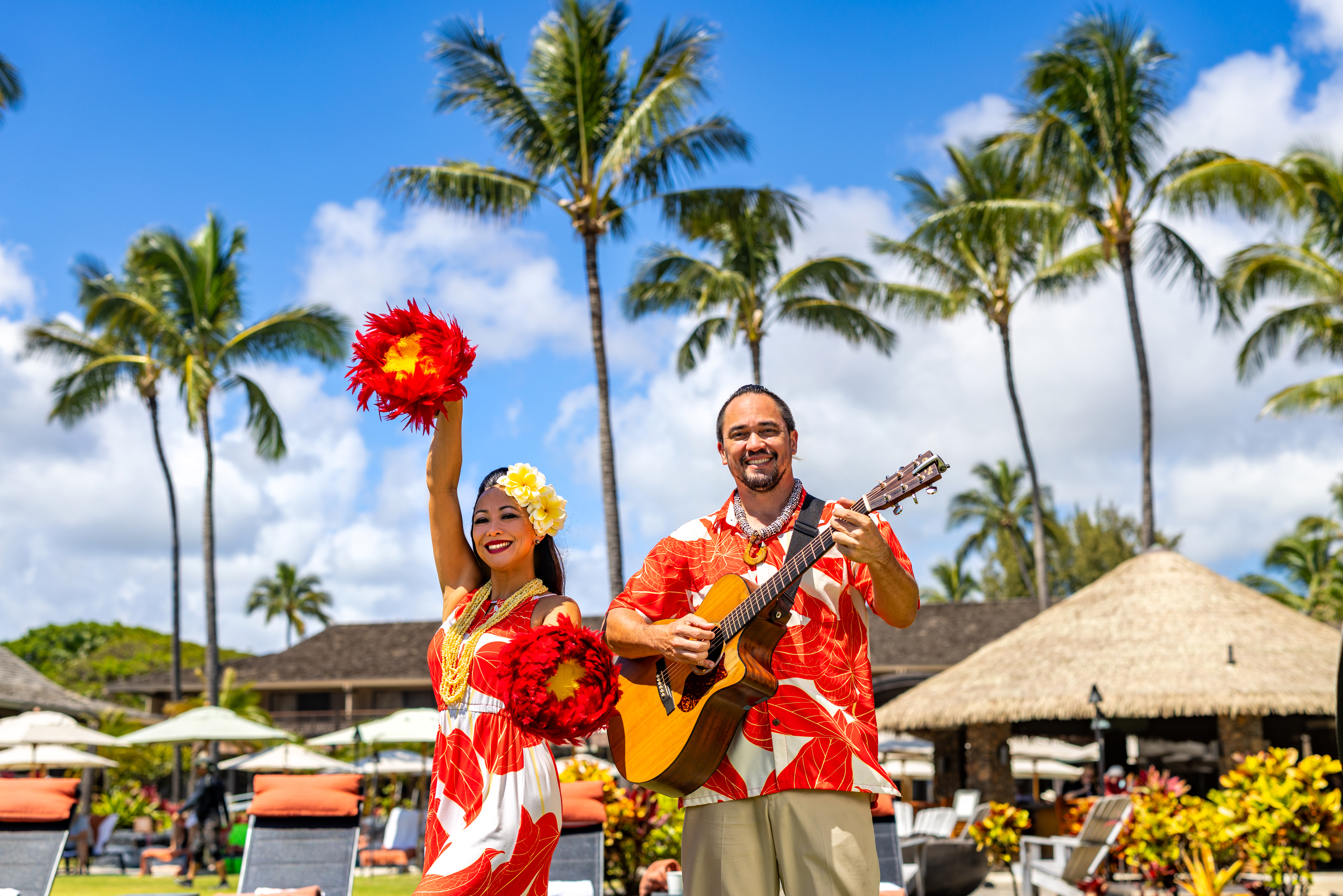 A woman in a red and white dress holds red feathered pom-poms while a man plays guitar; both stand outdoors with palm trees and thatched huts in the background.