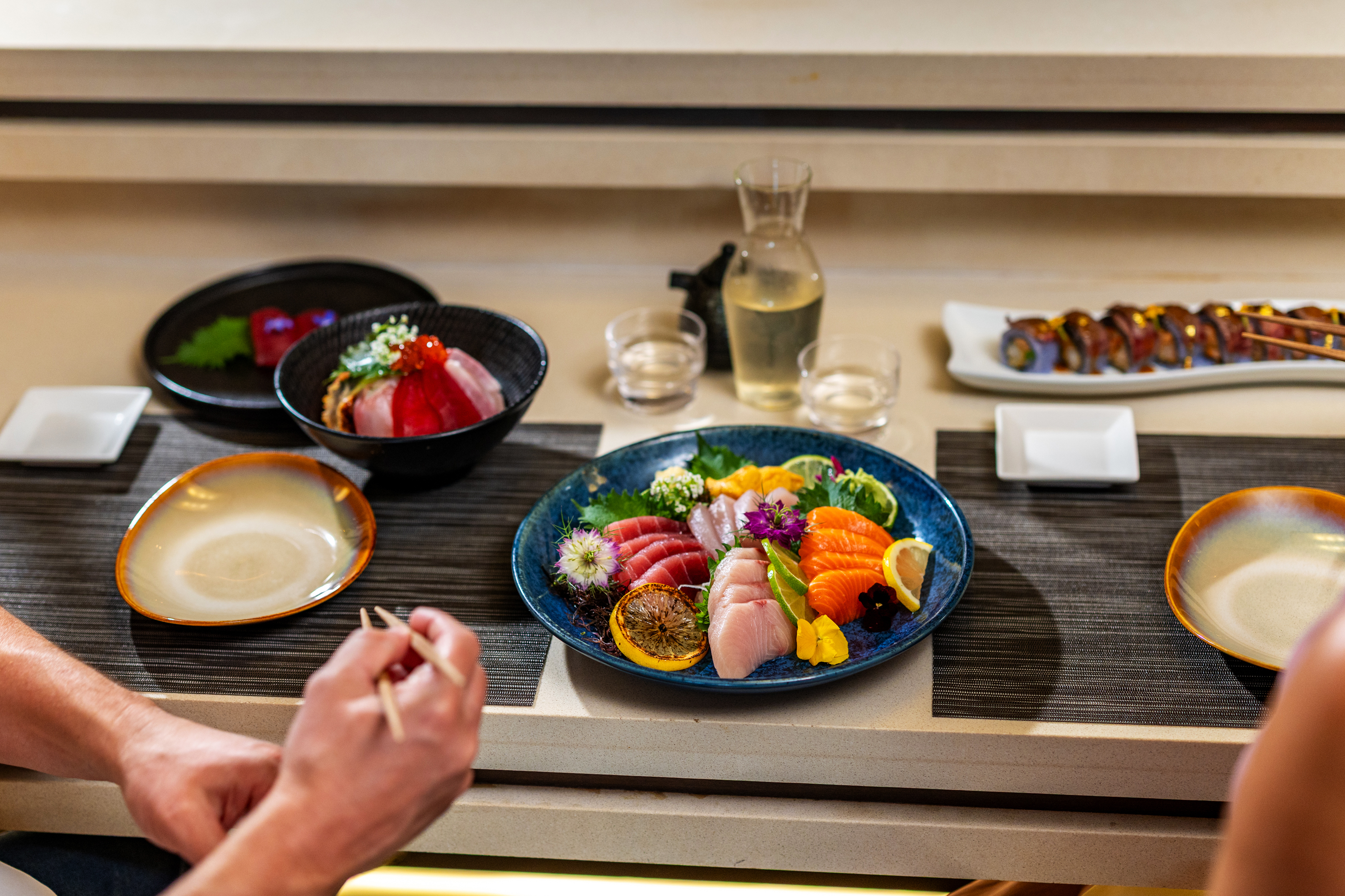 Two people dining at a table with assorted sashimi, a bowl of rice topped with fish, and a sake carafe with cups.