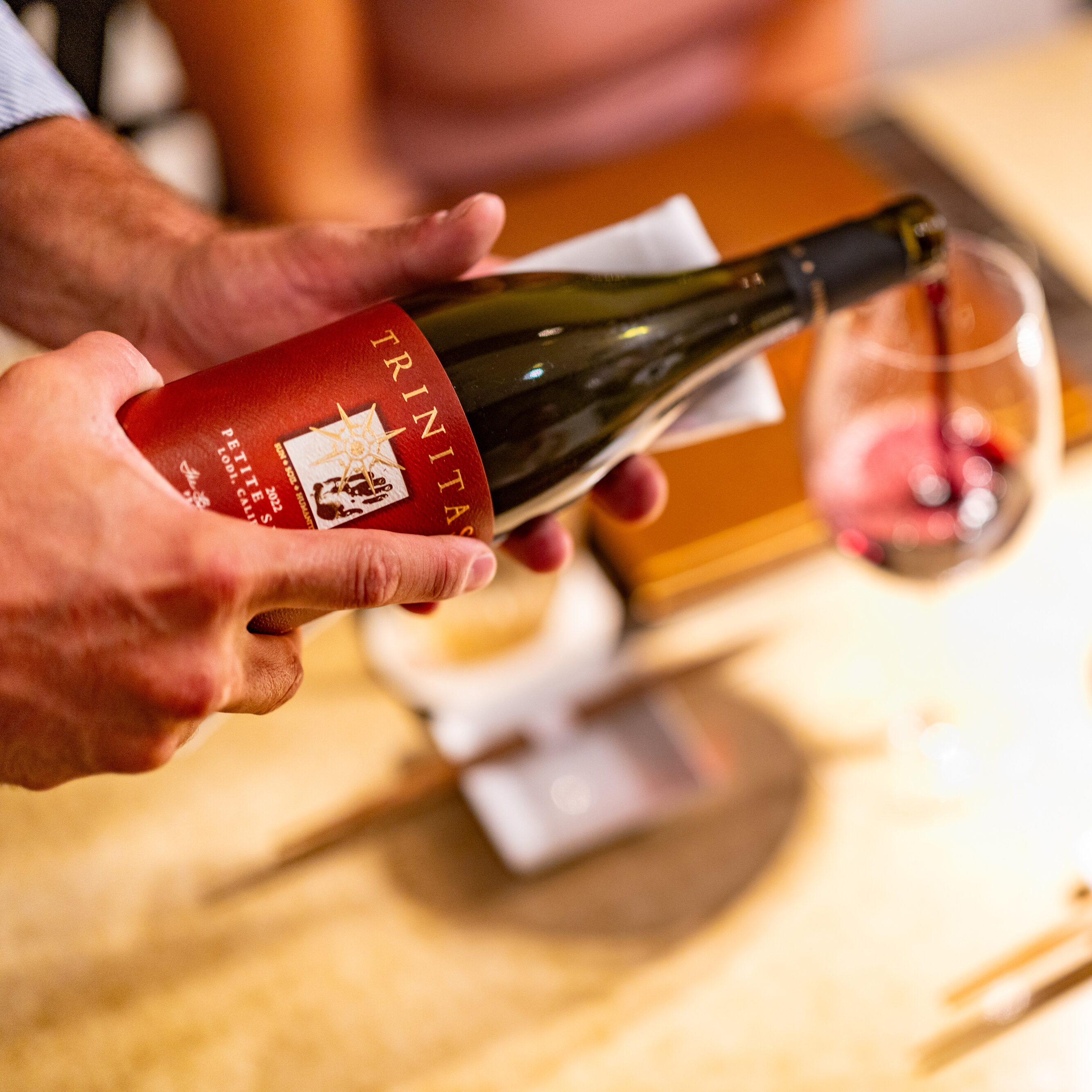 A person pours red wine from a Trinitas bottle into a glass on a dining table set for private dining, complete with utensils, a lamp, and hints of Hawaiian cuisine.