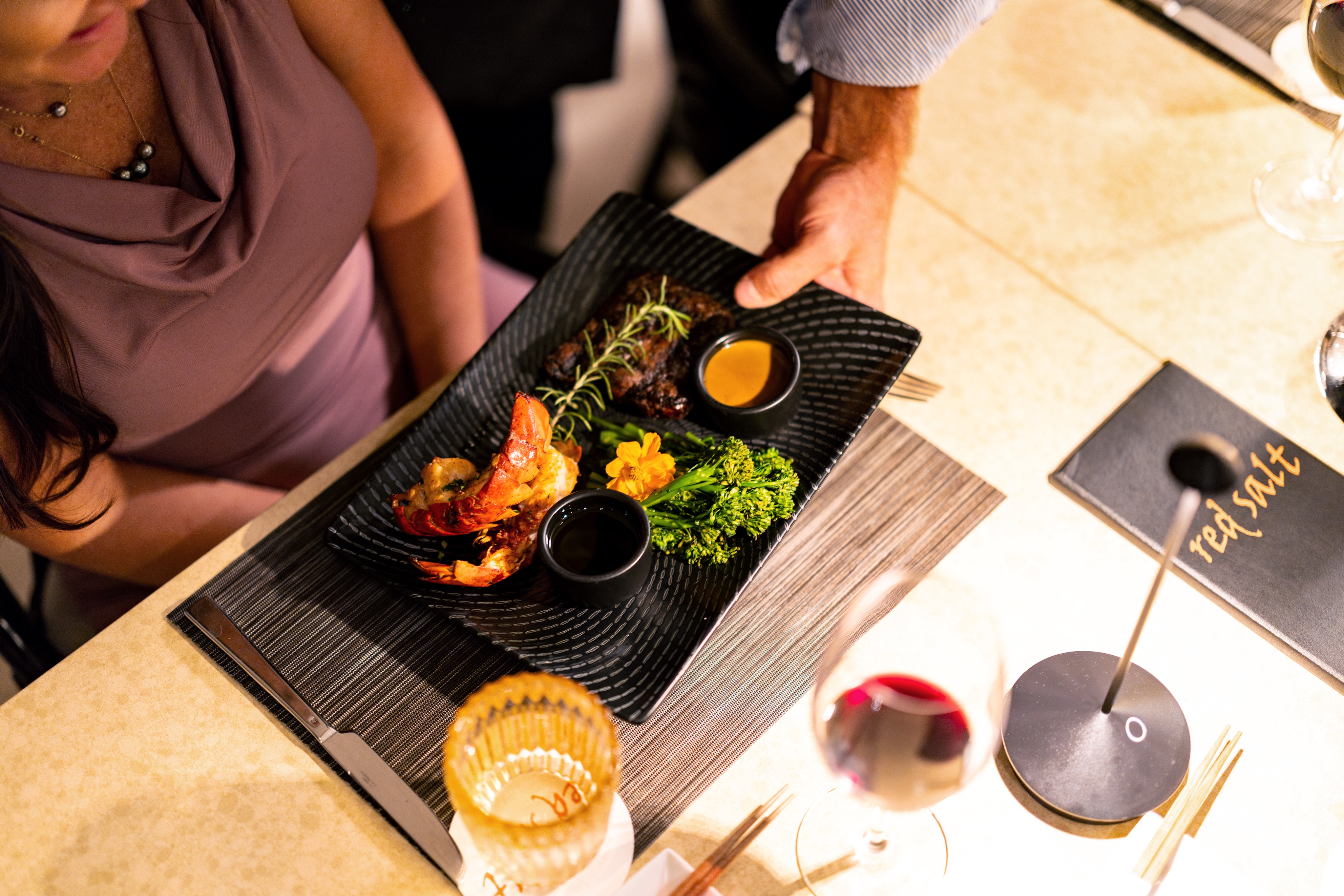 A person serves a black plate with shrimp, greens, and dipping sauce to a woman at a restaurant table set with wine, water, and menus.