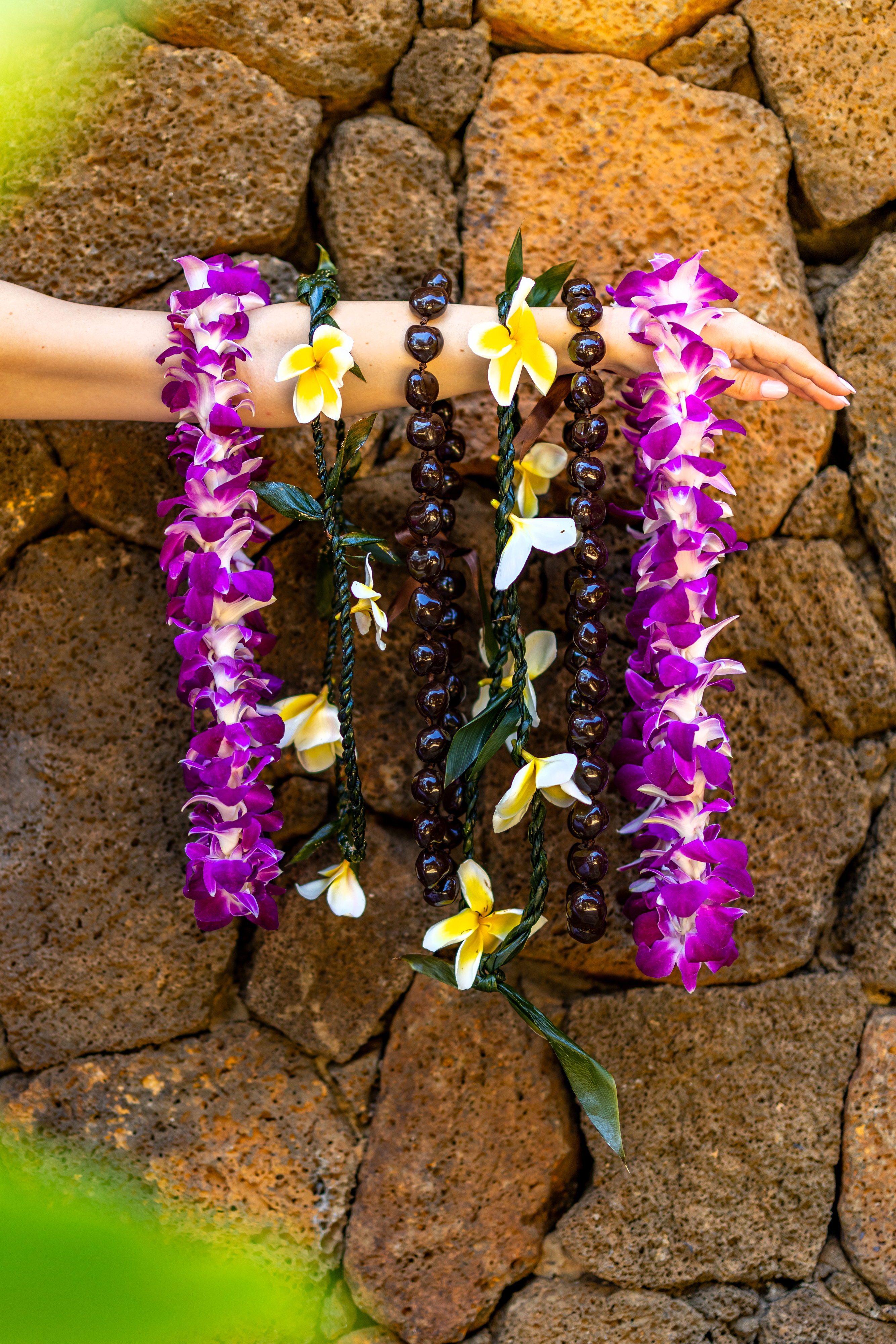 An outstretched arm displays several flower and kukui nut leis against a background of textured brown stone.