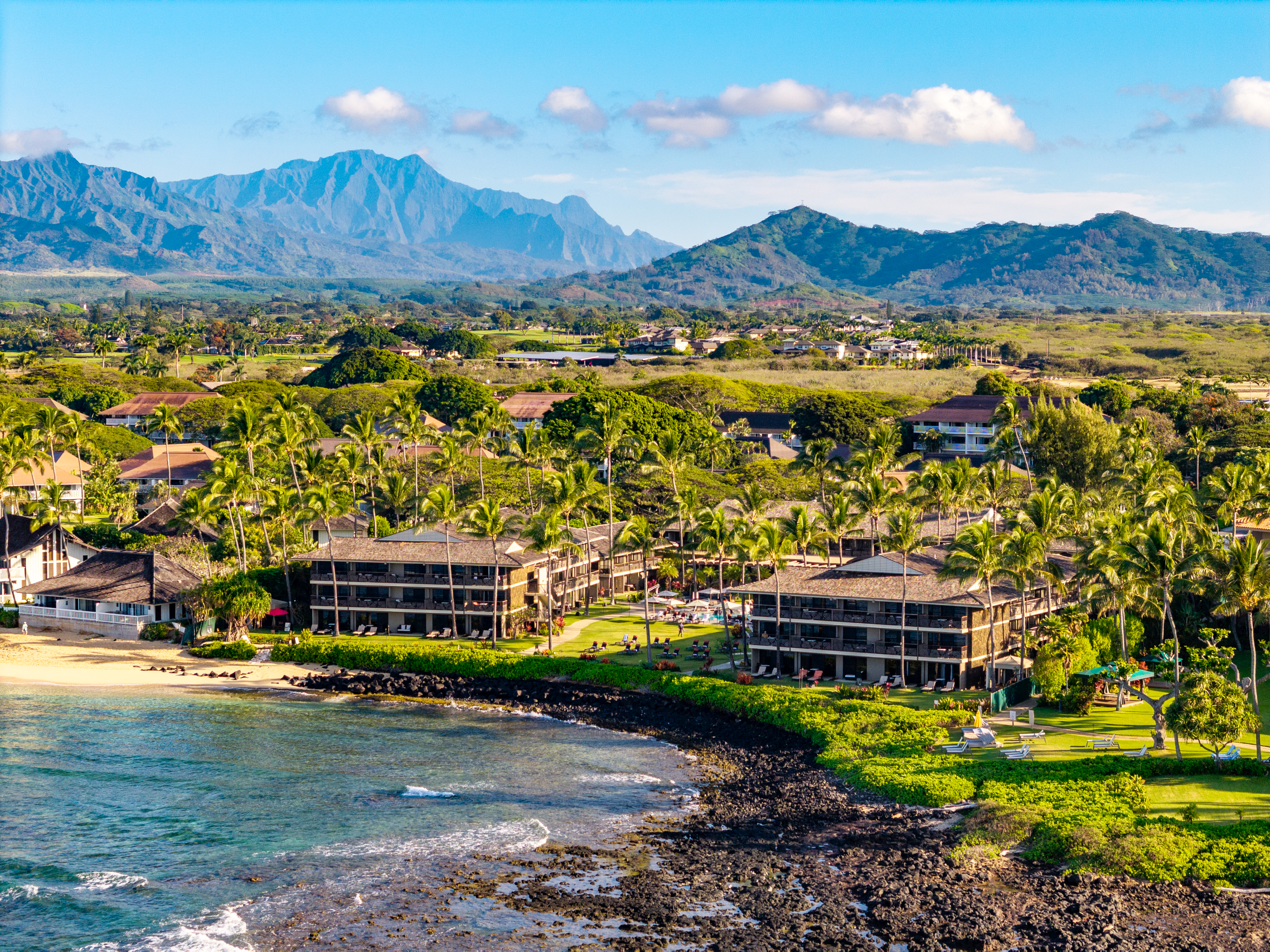 Aerial view of beachfront condos, palm trees, and rocky shoreline with mountains in the background under a blue sky.