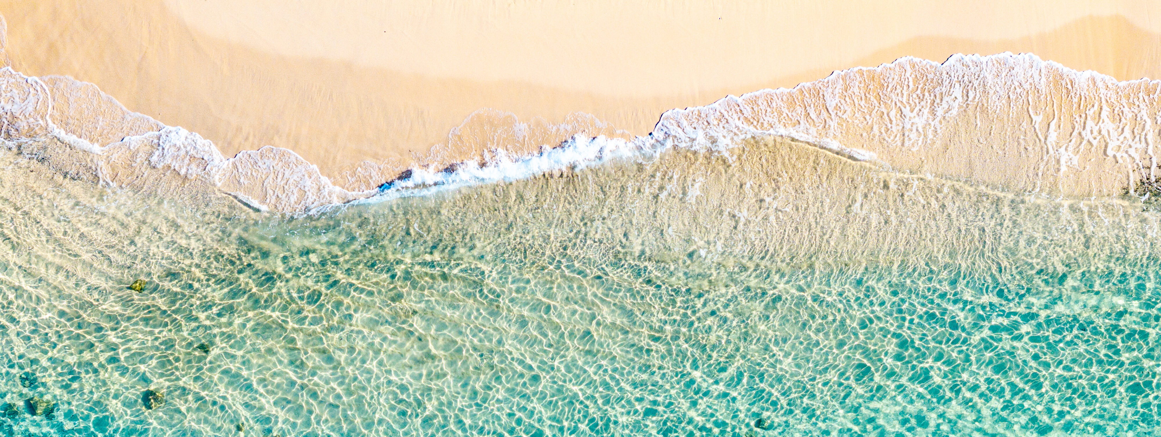 Aerial view of a sandy beach with clear turquoise water, gentle waves, and a visible underwater coral or rock formation near the shoreline.