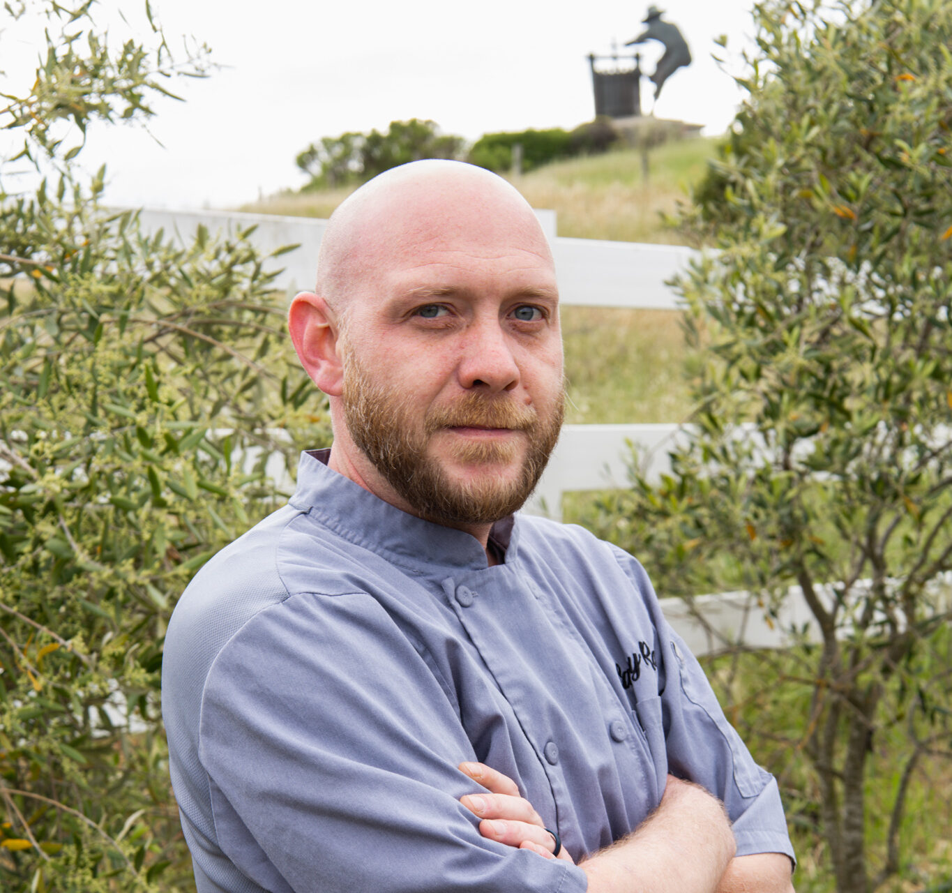 A bald man with a beard wearing a gray chef's coat stands outdoors with arms crossed, surrounded by greenery and a white fence in the background.