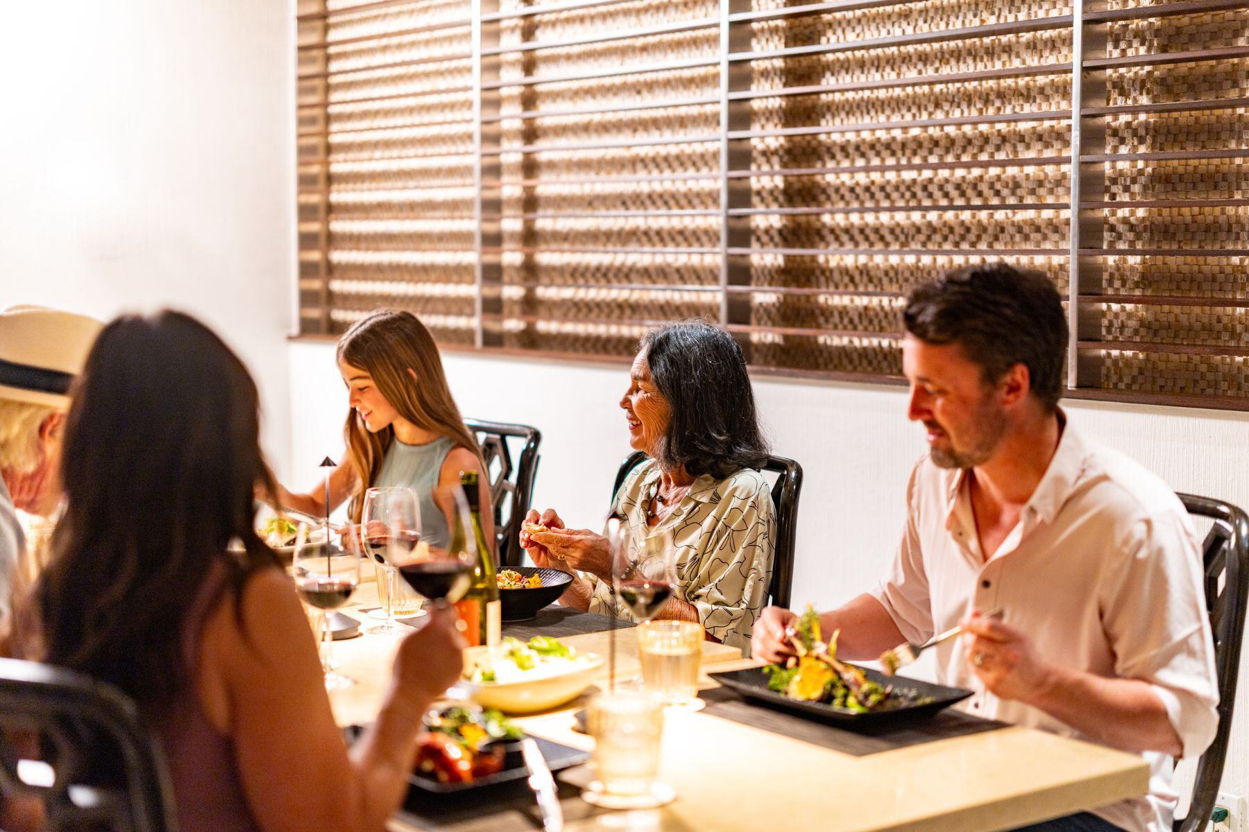 Five people sit around a dining table, enjoying Thanksgiving Dinner and drinking wine in a well-lit restaurant setting.