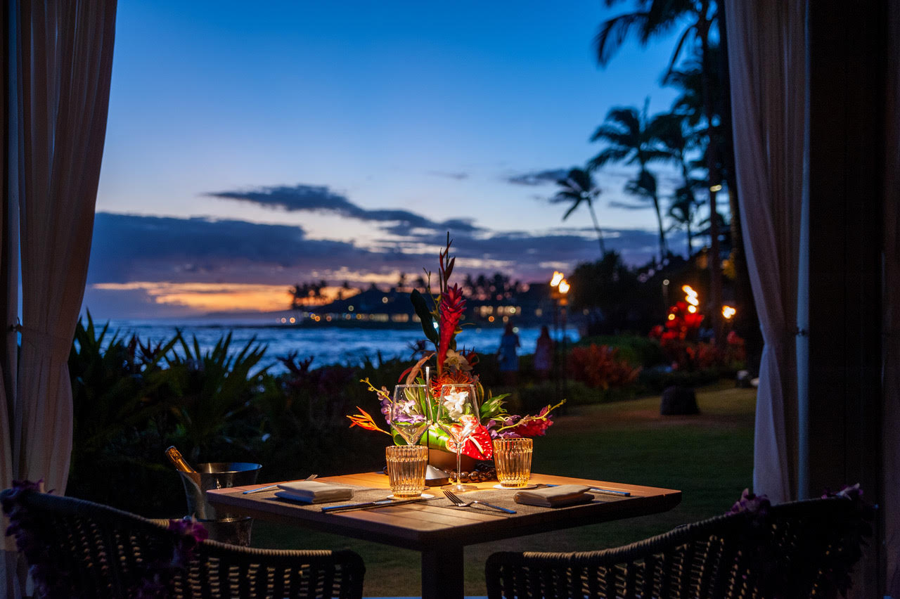 A small dinner table for two with glasses, plates, and a flower centerpiece overlooks a tropical garden and ocean at sunset—perfect for intimate Cabana Dinners.
