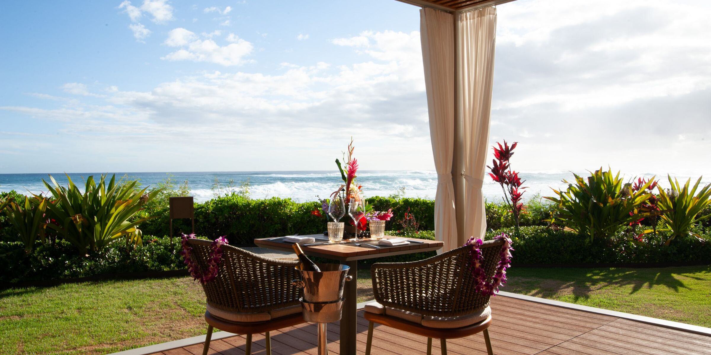 A table for two set for dining on a patio overlooking the ocean, surrounded by tropical plants and clear skies—perfect for romantic Cabana Dinners with serene seas in the background.