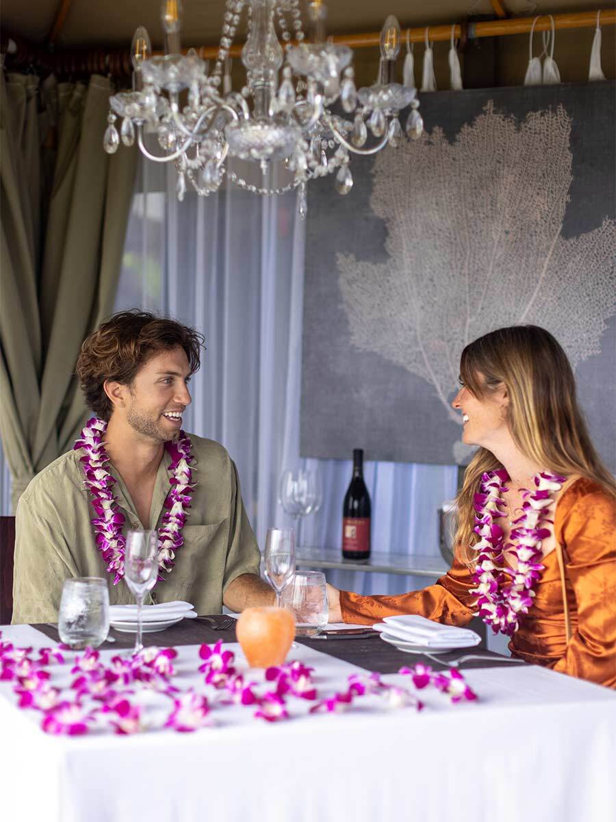 A man and a woman wearing flower leis sit at a table decorated with flower petals, enjoying a meal together in a well-lit, elegant setting—perfect for unforgettable Cabana Dinners.