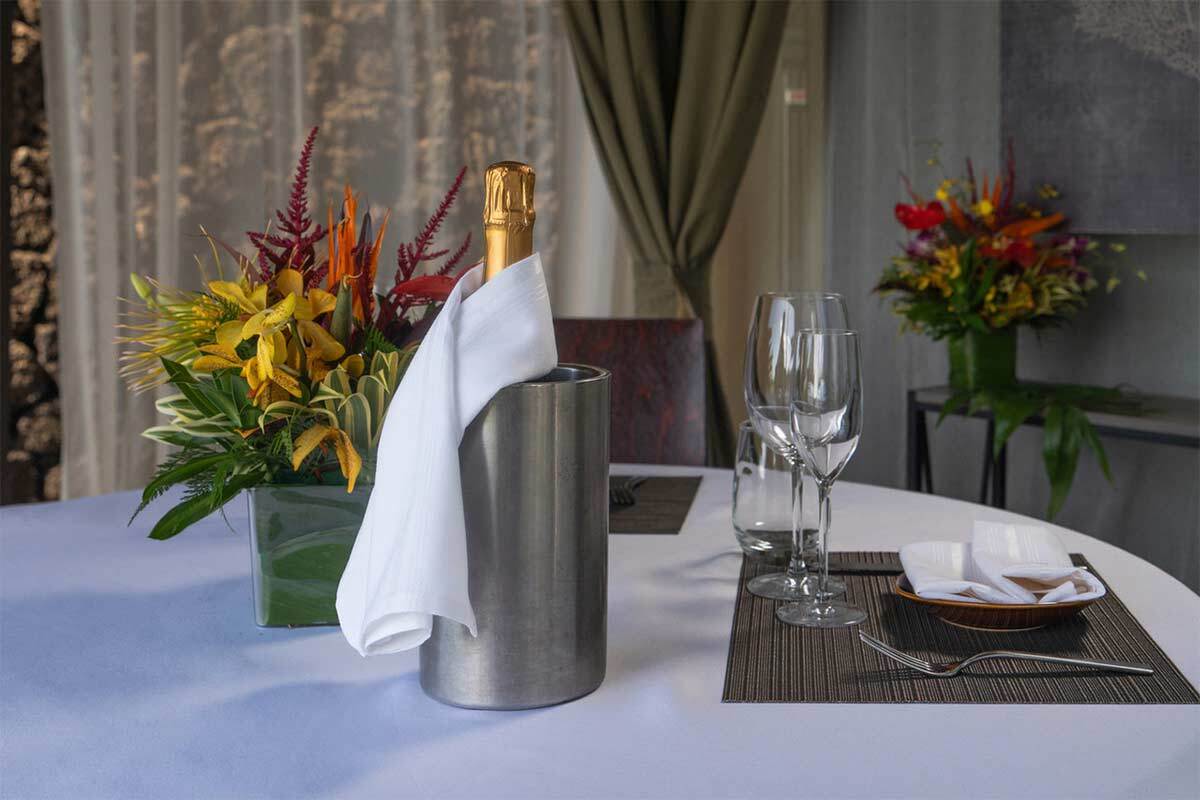 A table set for Cabana Dinners features a champagne bottle in an ice bucket, a white napkin, two glasses, plates, and a beautiful flower arrangement centerpiece.