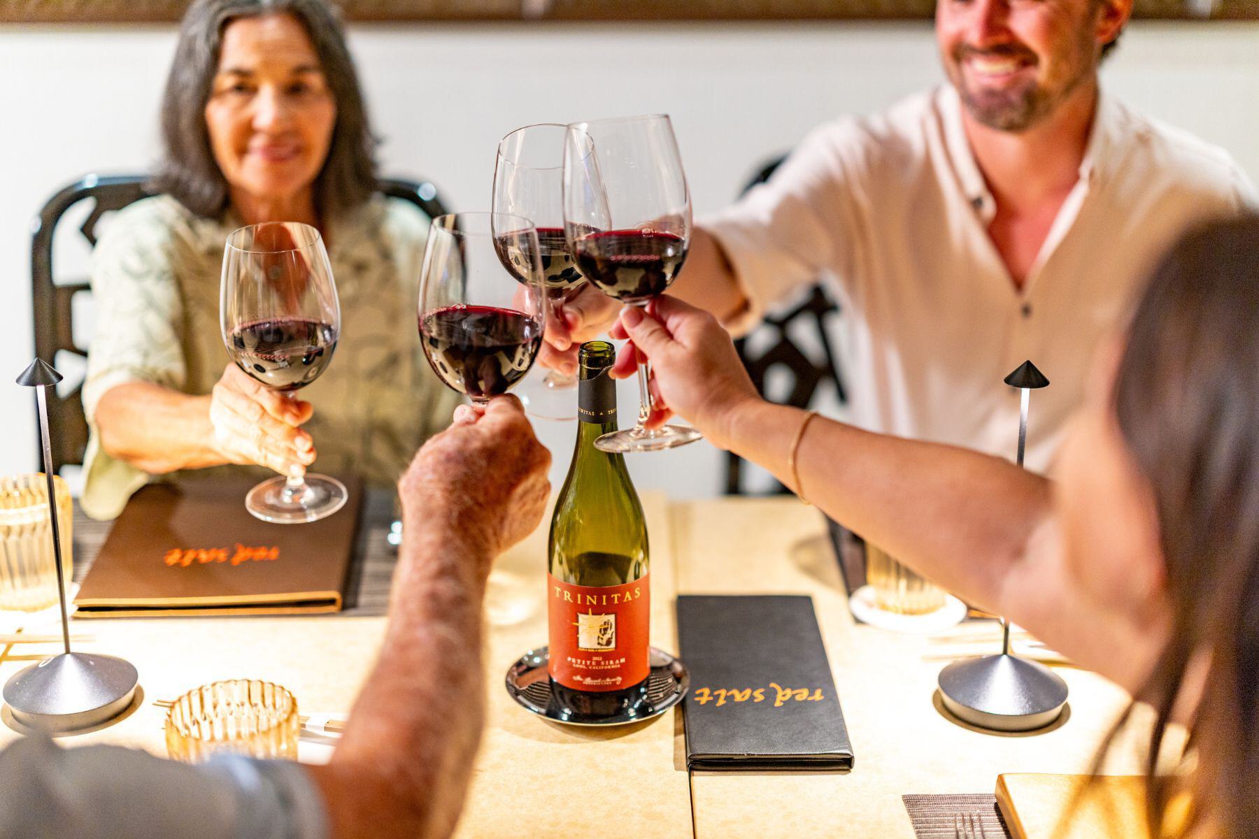 Four people clink glasses of red wine over a restaurant table set with menus, a bottle of Trinitas wine, and glasses—celebrating with friends and savoring every pour. Don’t miss the Black Friday offer on your favorite wine!.