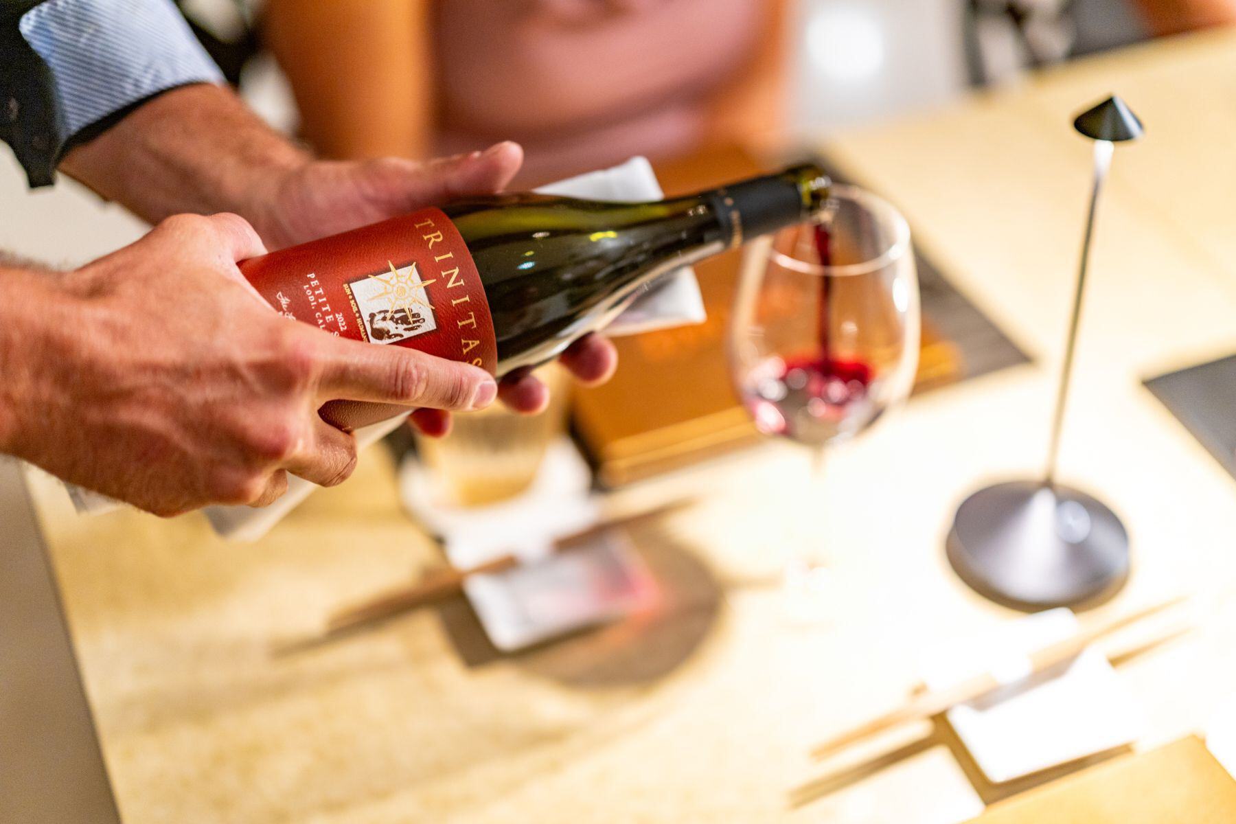 A person pours red wine from a bottle into a glass on a restaurant table set with chopsticks and napkins, creating the perfect setting for a romantic Valentine's Day dinner.