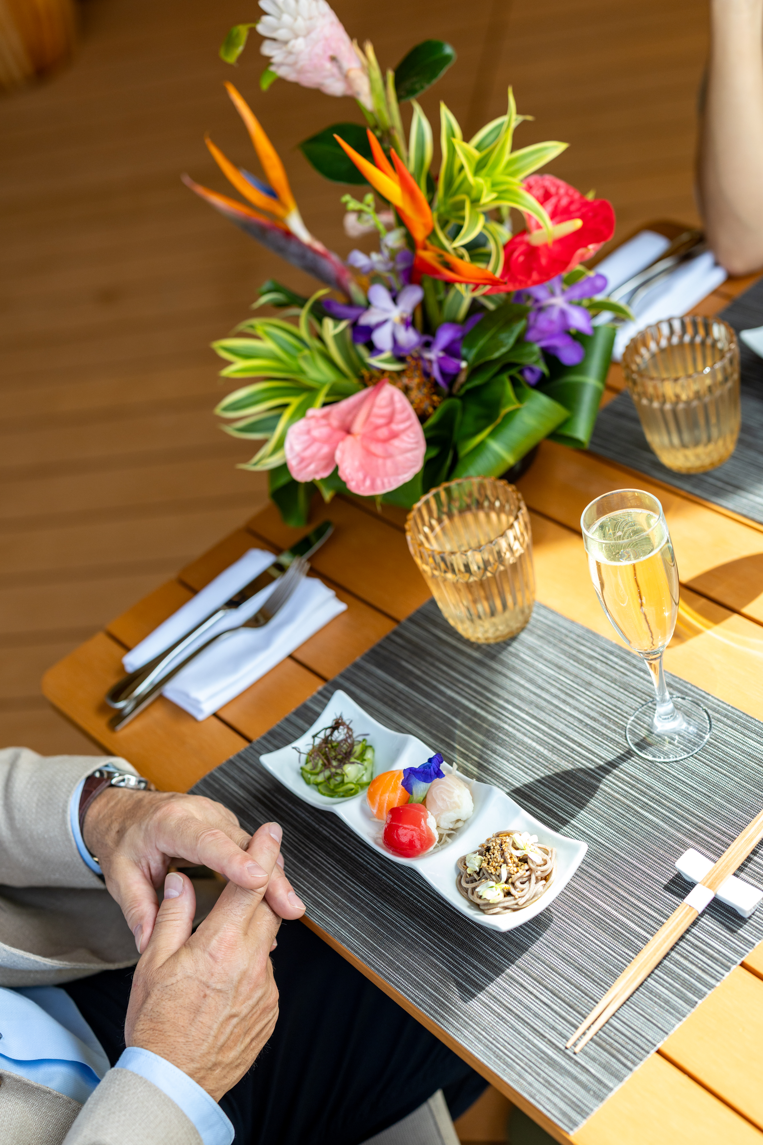 A person sits at a table set with small plates of seafood, a glass of champagne, cutlery, chopsticks, and a colorful flower arrangement, savoring the relaxed elegance of Cabana Dinners.