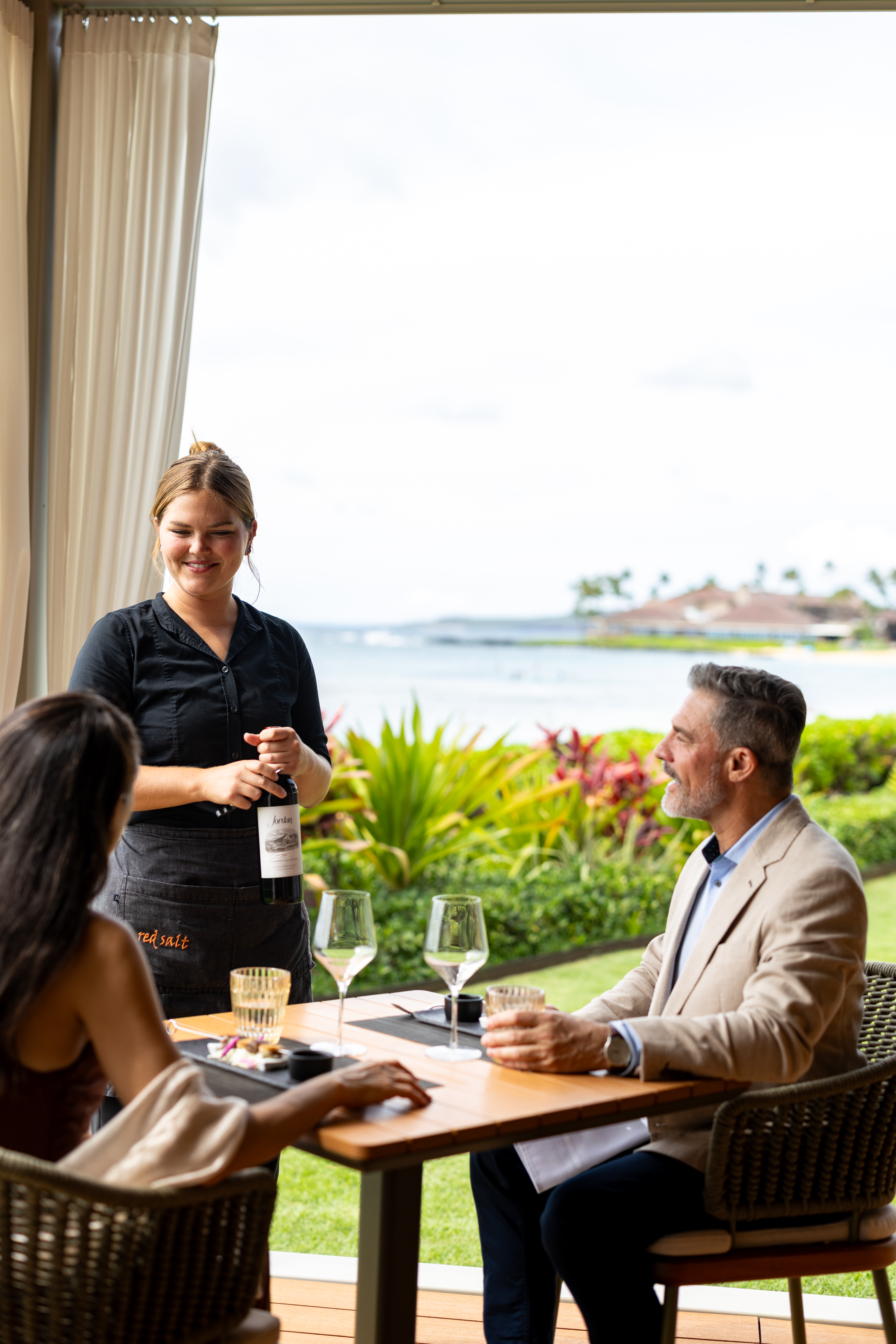 A server presents a bottle of wine to a seated couple enjoying Cabana Dinners at an outdoor restaurant with a scenic ocean view.