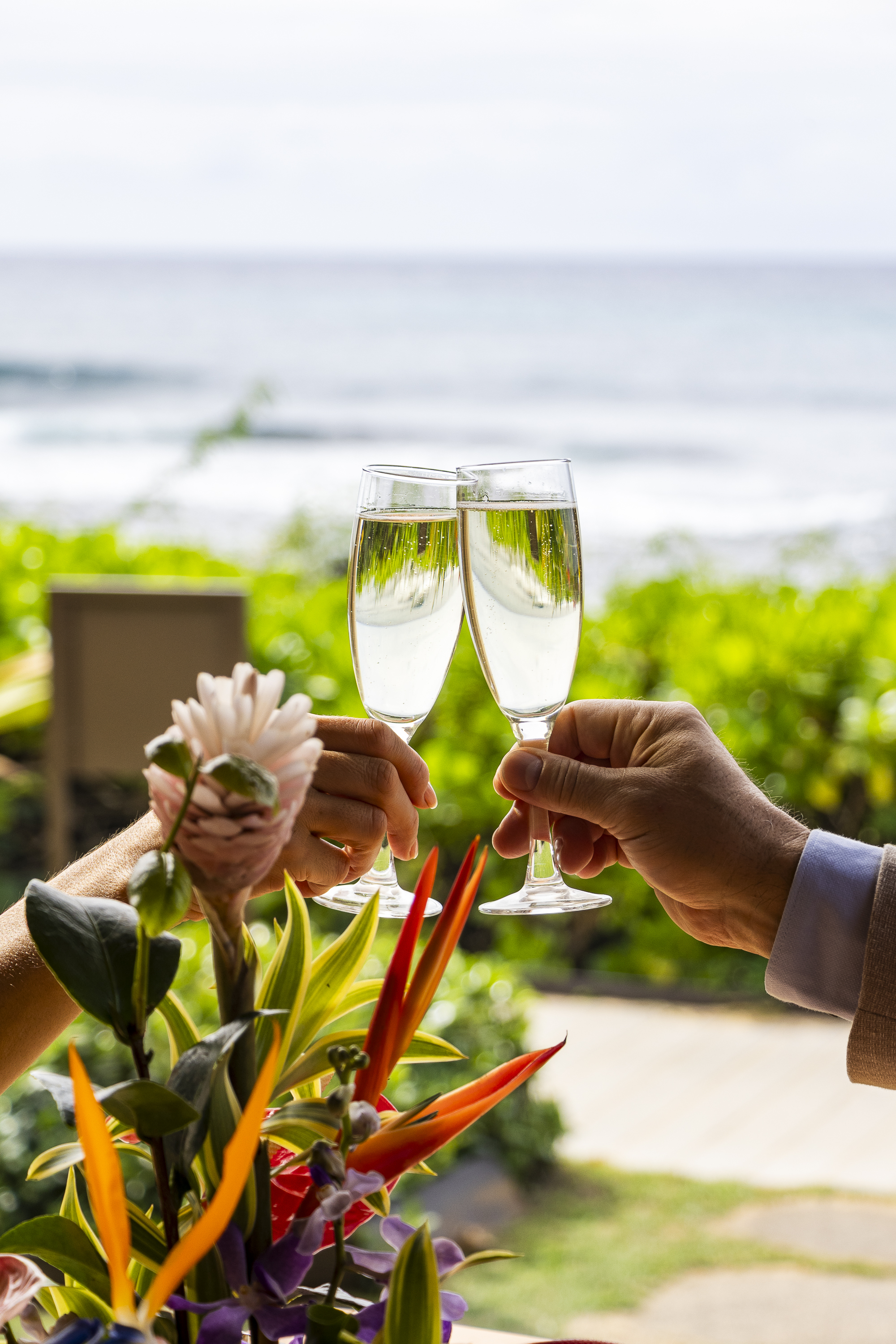 Two people clink champagne glasses near a bouquet of tropical flowers, enjoying Cabana Dinners with a blurred ocean and lush greenery in the background.