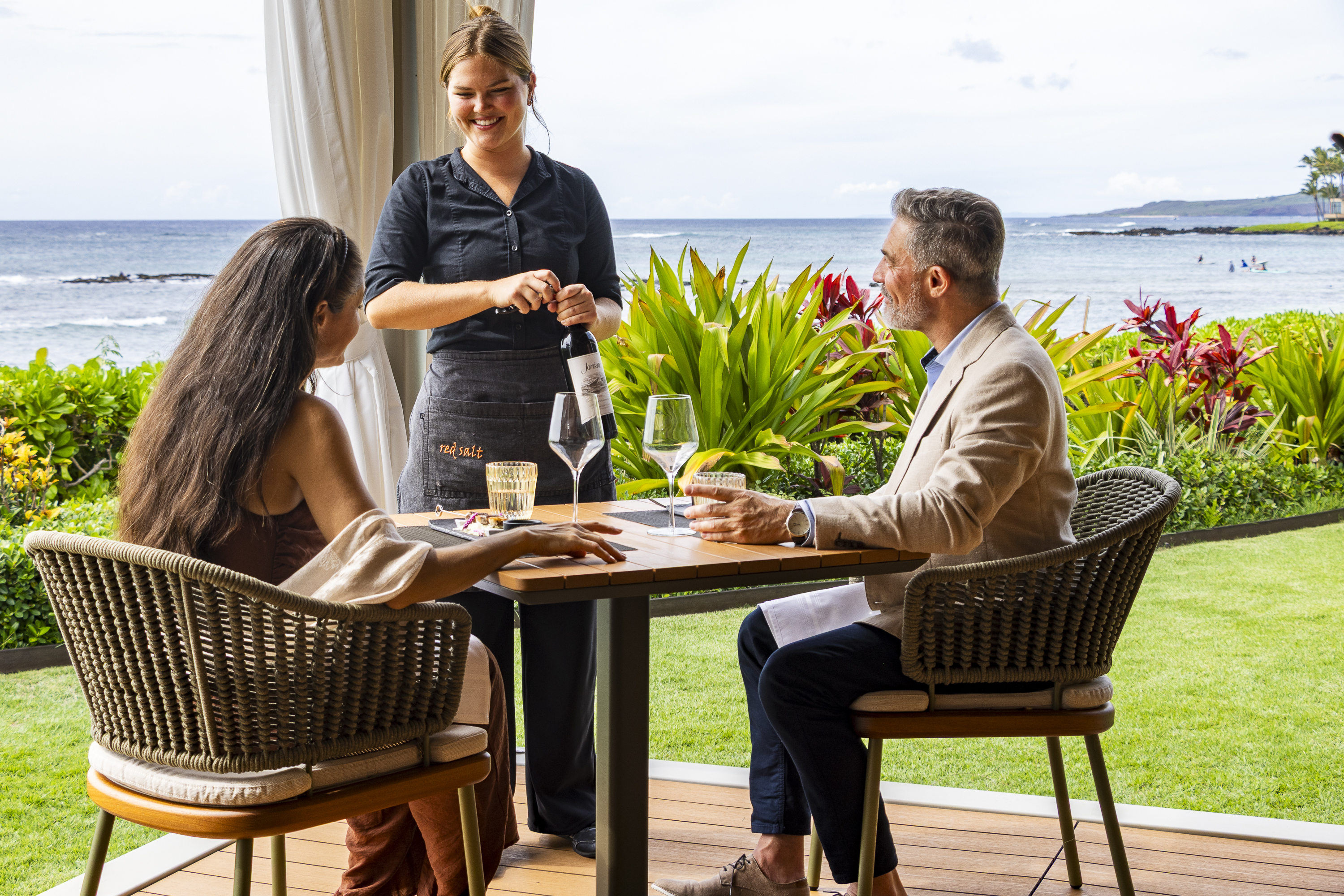 A server opens a wine bottle at an outdoor restaurant table for a seated man and woman during their Cabana Dinners, with ocean and greenery in the background.