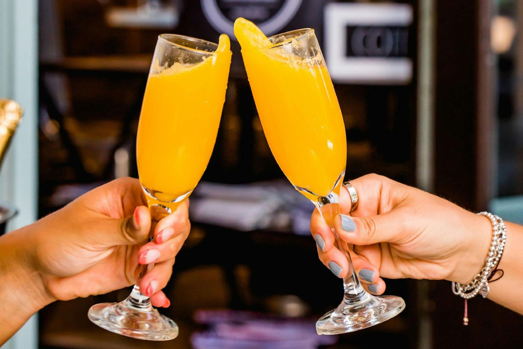 Two hands holding champagne flutes filled with an orange beverage, clinking glasses together in a toast—perfect for Mother's Day Brunch at Red Salt.