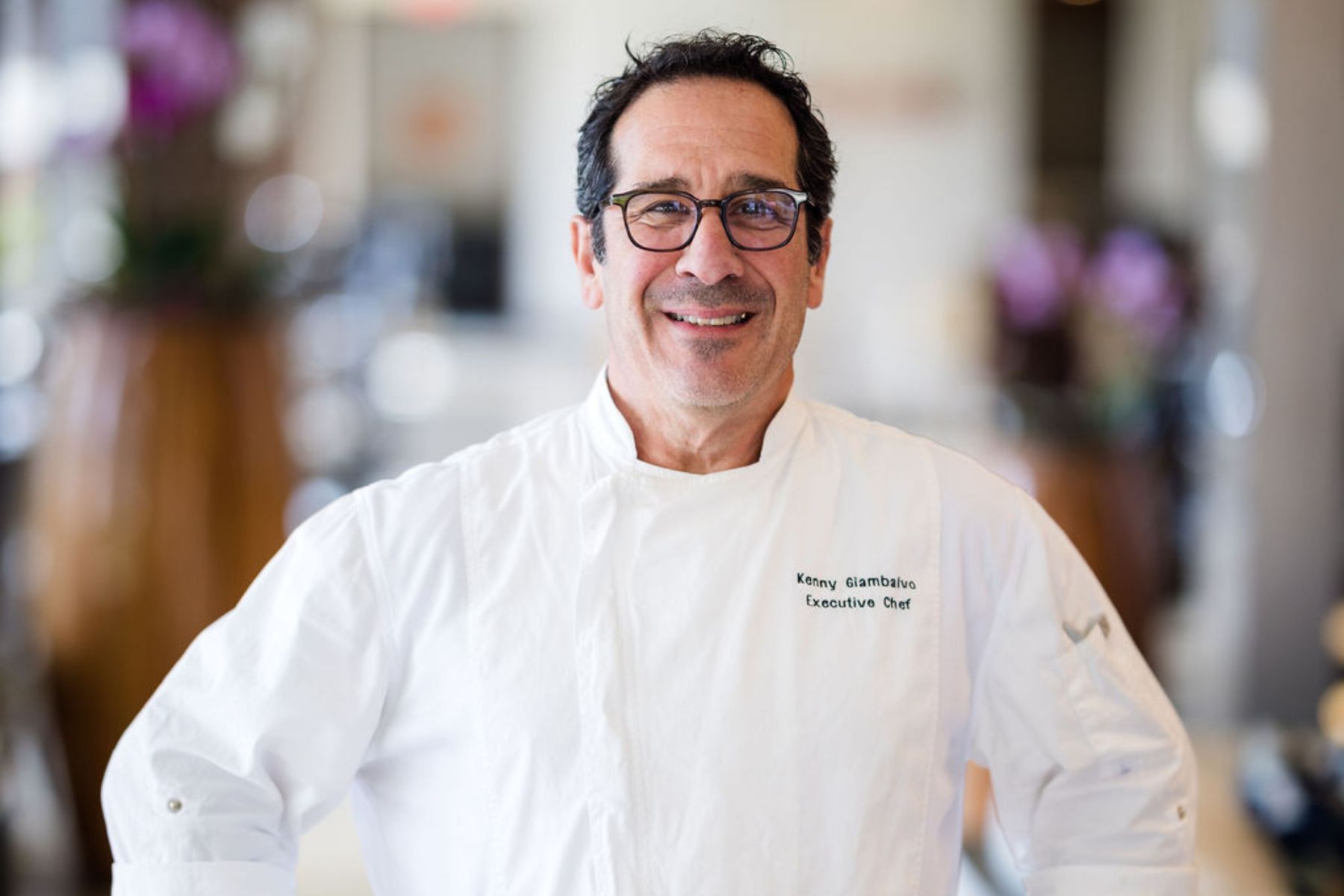 A man wearing glasses and a white chef’s coat stands indoors, smiling at the camera. The chef’s coat reads “Kenny Giambalvo, Executive Chef,” as he prepares for the Dinner Pa'ina Series.