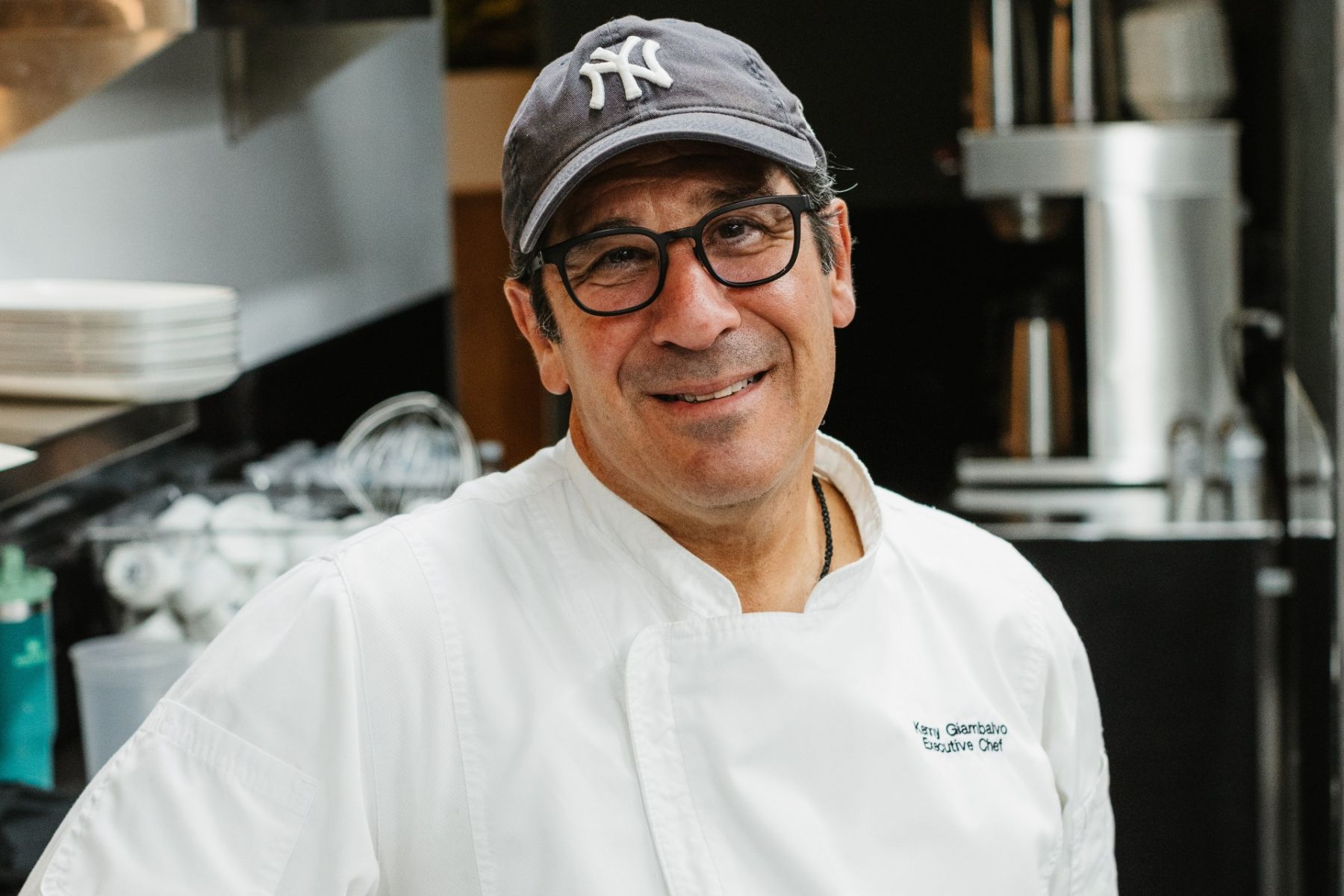 Chef Kenny Giambalvo, wearing glasses, a baseball cap, and a white chef’s coat, stands and smiles in a commercial kitchen during the Dinner Pa'ina Series.