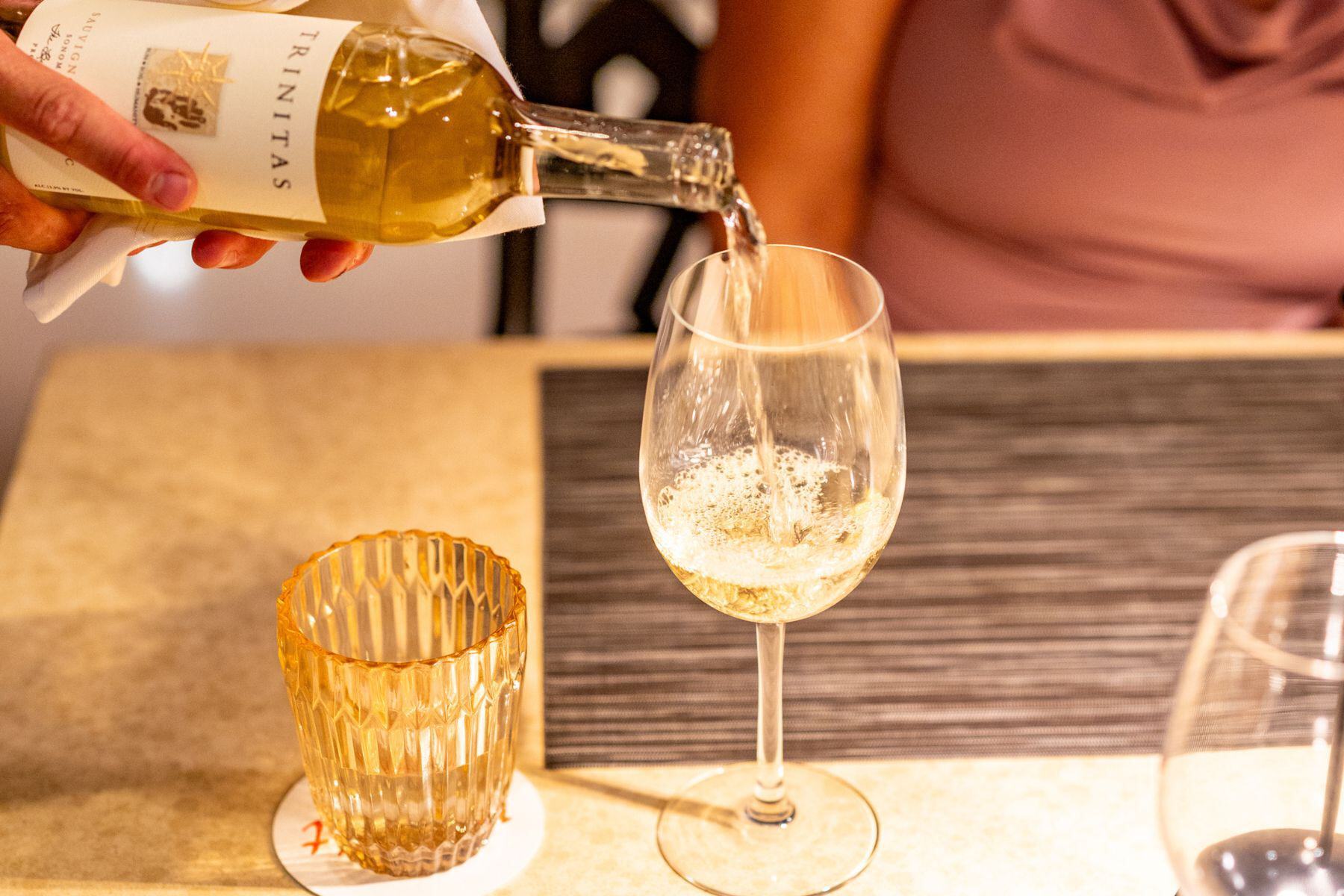 A hand pours white wine from a bottle into a glass during Pau Hana, with another glass, a coaster, and a placemat arranged on the table for this relaxing happy hour moment.