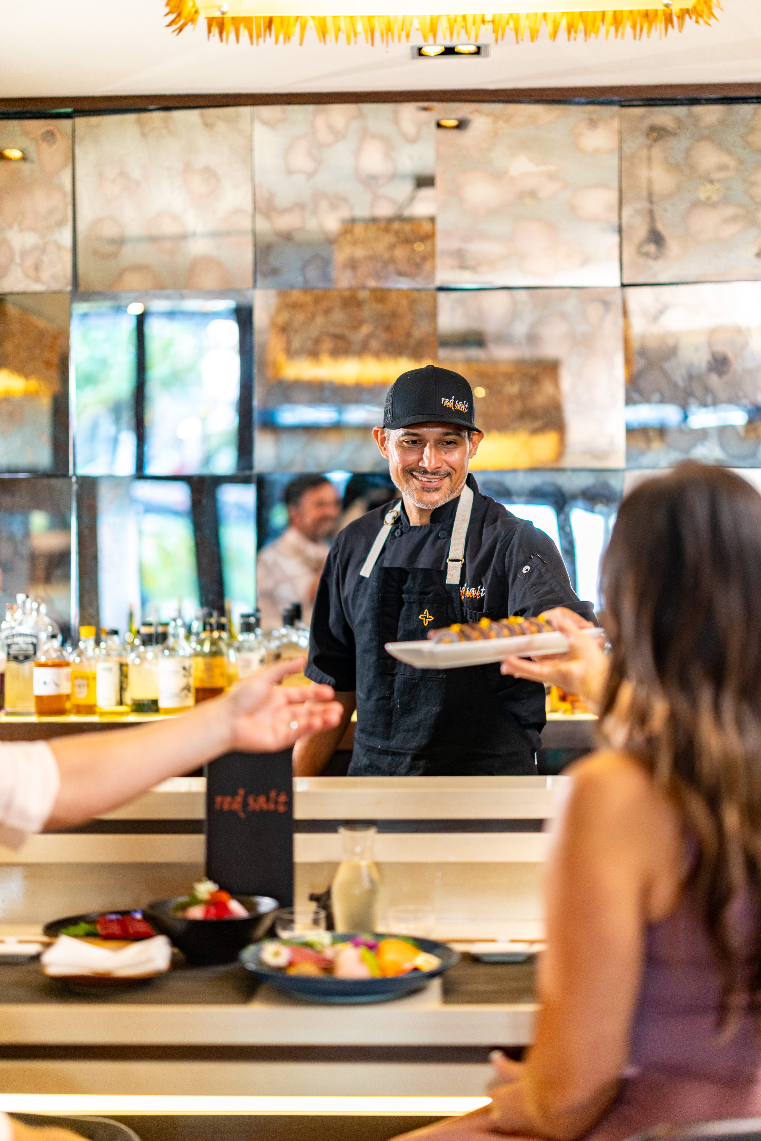 A chef in a black uniform and cap smiles while handing a plate of food to a seated customer at a modern restaurant bar, celebrating National Hand Roll Day with fresh, creative dishes.