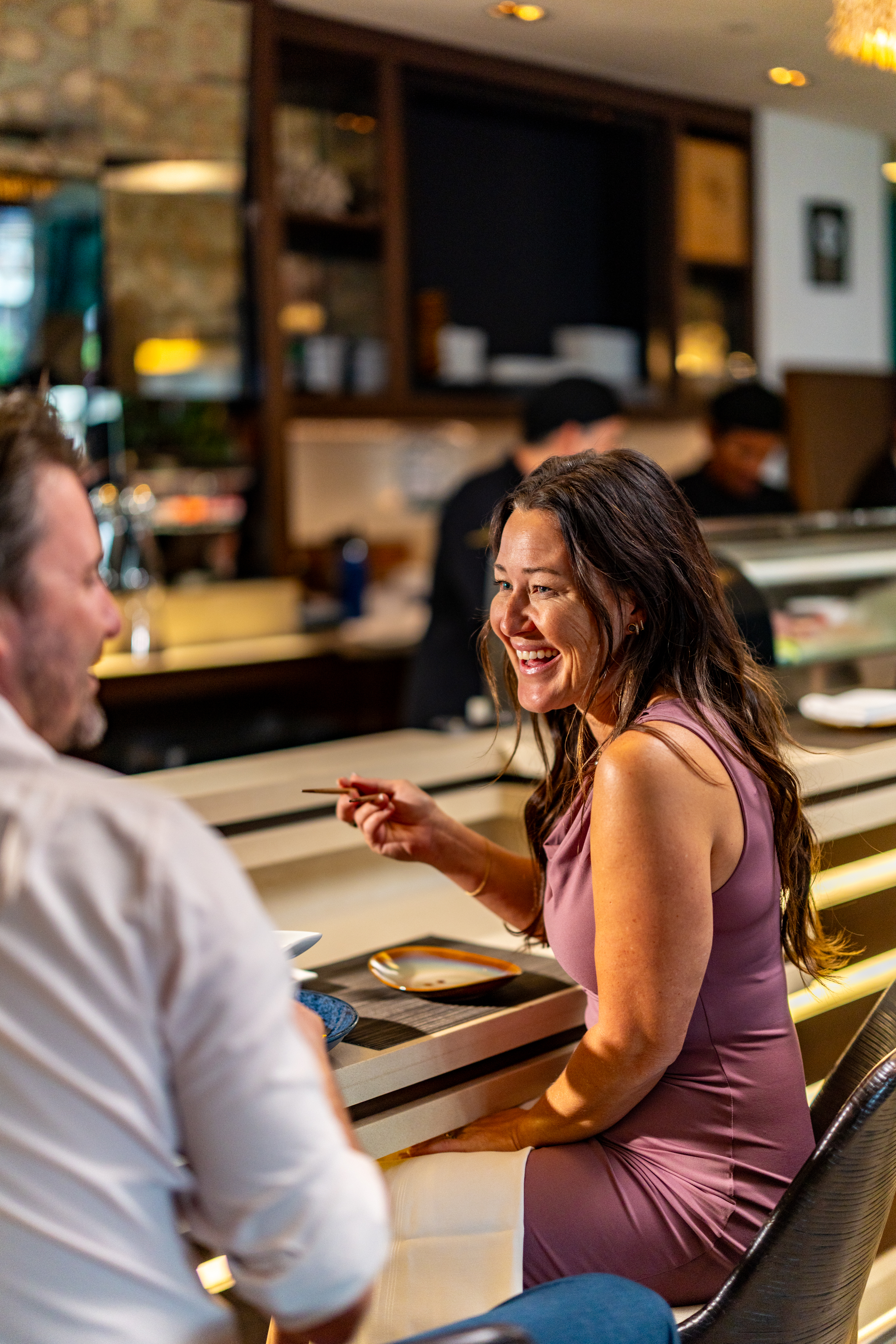 A woman in a sleeveless lavender dress smiles and holds chopsticks, celebrating National Hand Roll Day as she sits at a restaurant counter across from a man. Chefs work behind the counter in the background.