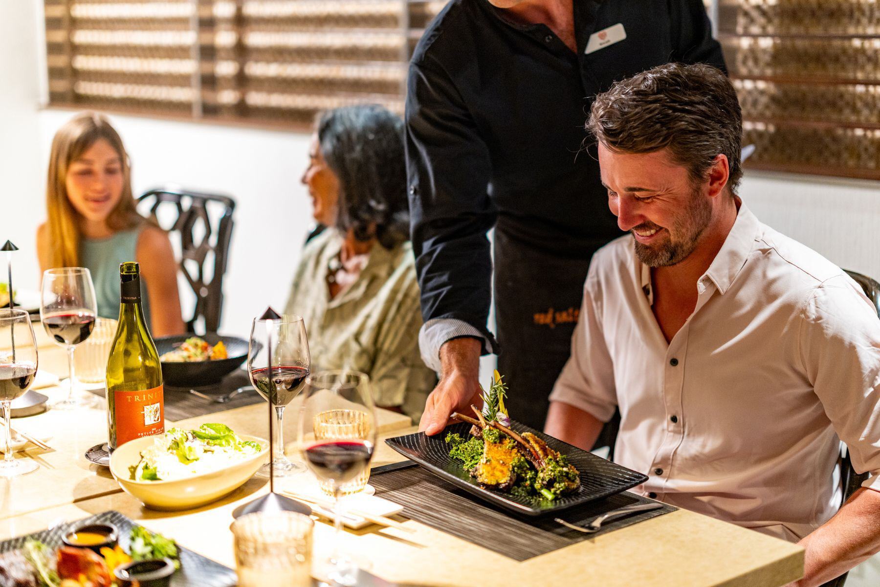 A server presents a plated meal to a smiling man enjoying Father’s Day at Red Salt, seated at a restaurant table with three other people, wine glasses, and food dishes.