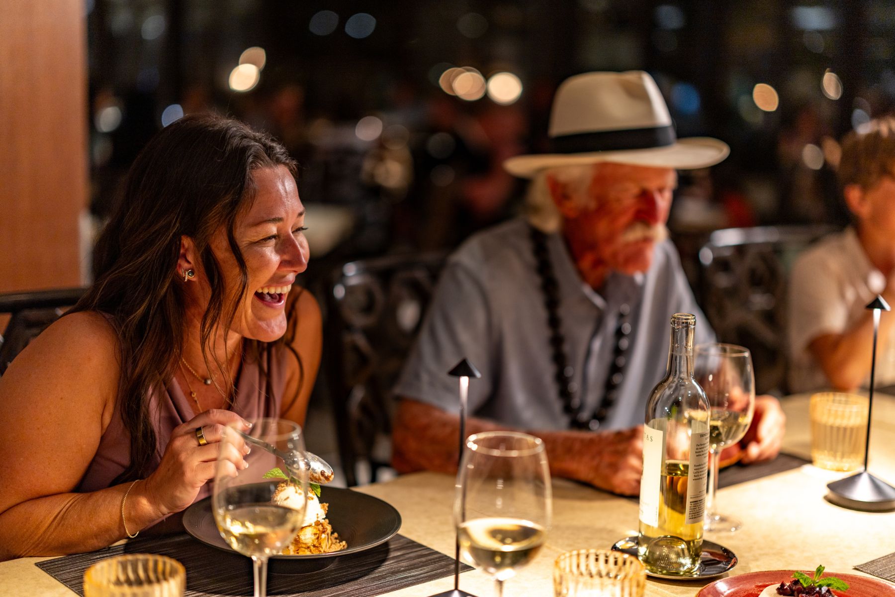 A woman laughs while eating a prix-fixe menu at a restaurant table with an older man in a white hat; wine glasses and plates are in front of them.