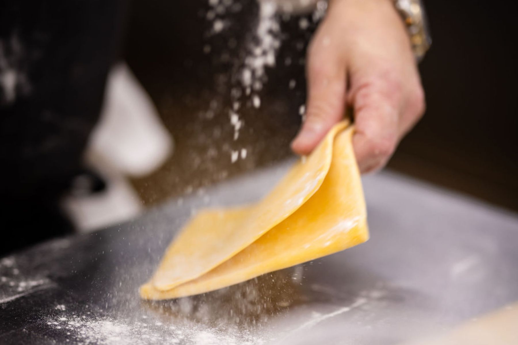 A hand folds a sheet of yellow pasta dough on a floured surface, with flour particles visible in the air—evoking the careful preparation behind every course of a prix-fixe menu.
